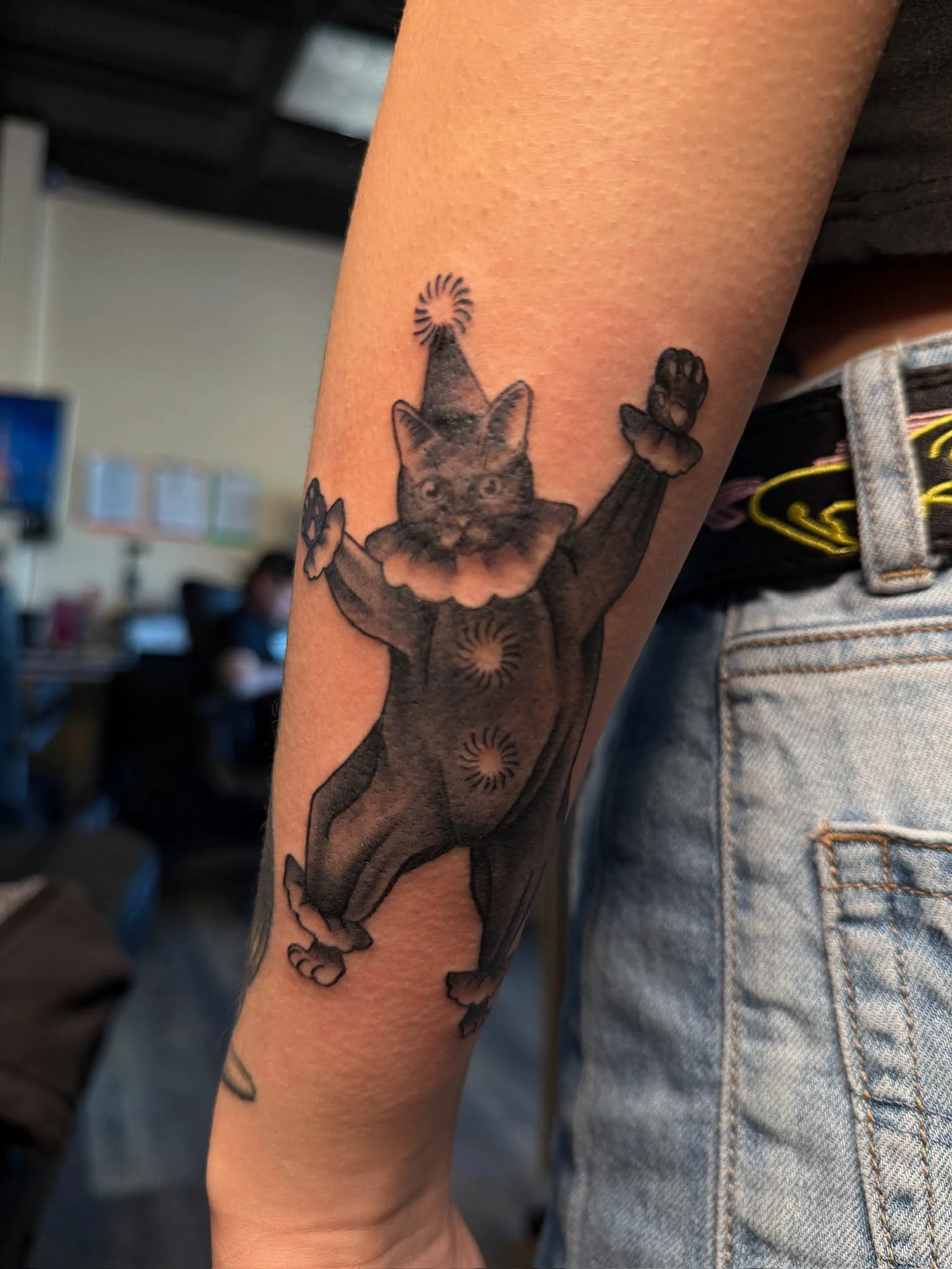 Tattoo of a cat dressed as a clown with a party hat, with its arms raised.