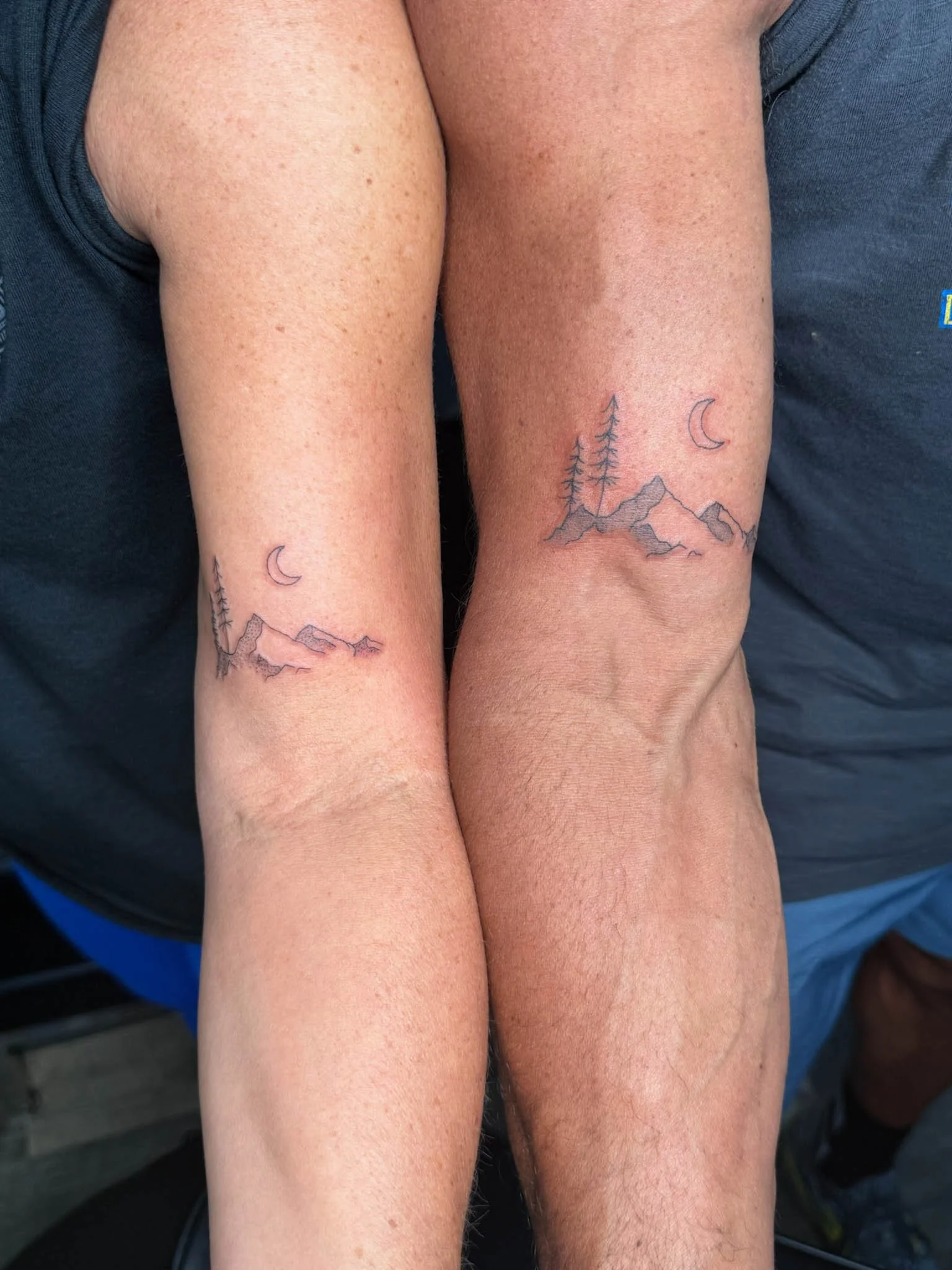 Two people with matching mountain and tree tattoo designs on their upper arms, featuring thin black outlines, a small crescent moon, and a mountain range, with slight variations in the tattoos.