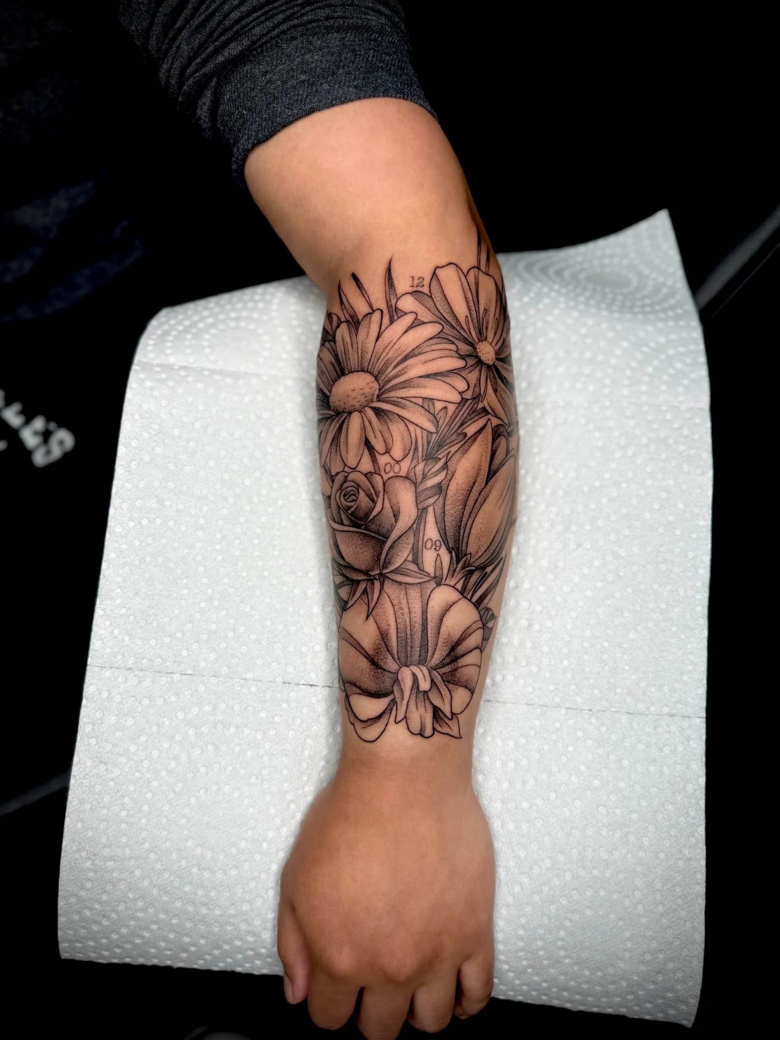 Black and gray floral tattoo on person's forearm, featuring daisies, roses, tulips, and various leaves.