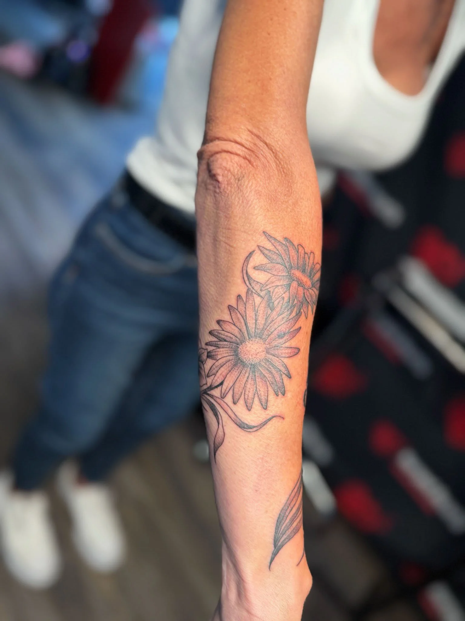 A close-up of a person's forearm with a tattoo of two large daisies and a long leaf in black ink.
