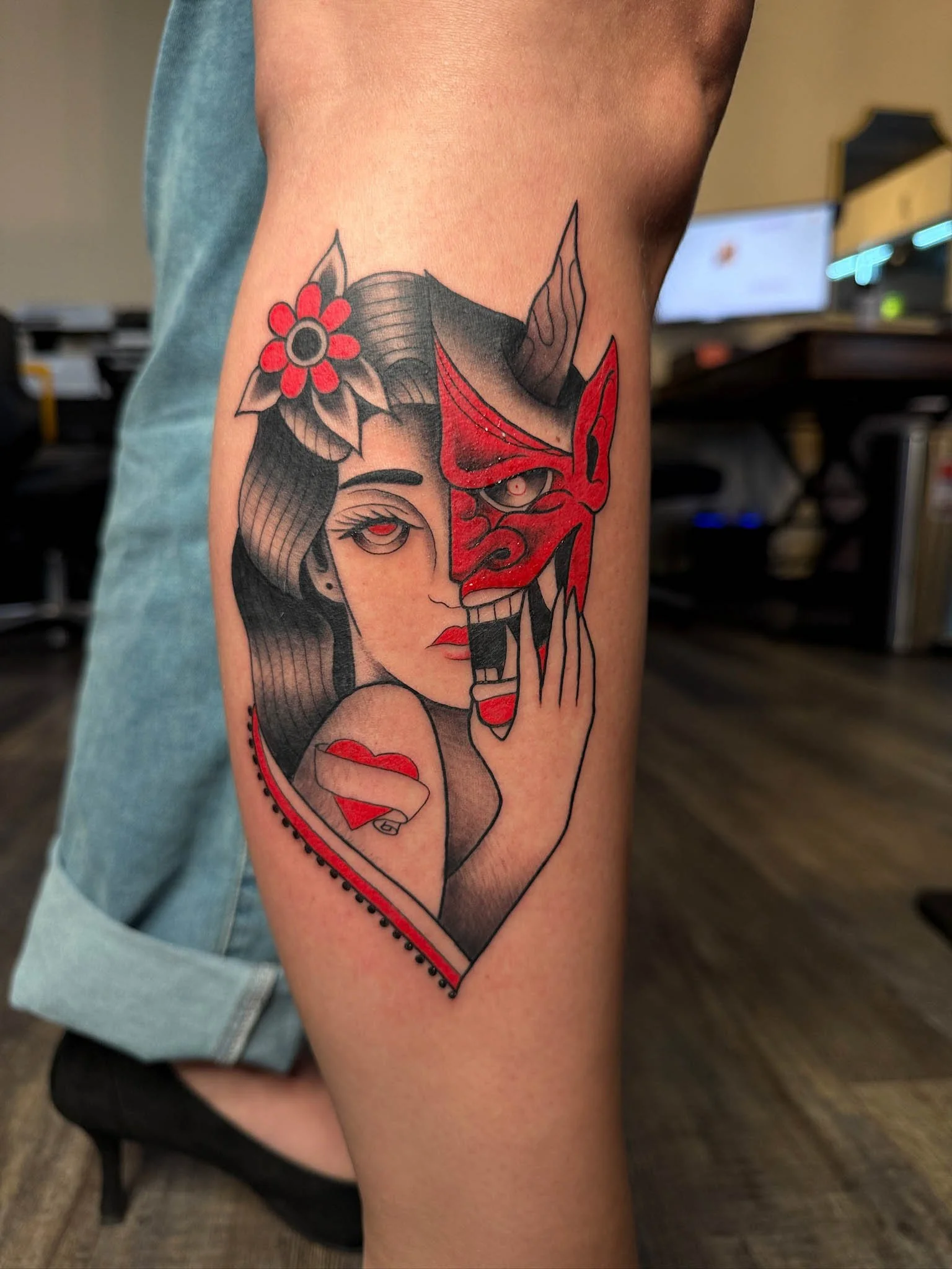 Tattoo of a woman with dark hair and a flower in her hair, holding a red mask, with her face split between a woman and a demon face, within a heart outline.