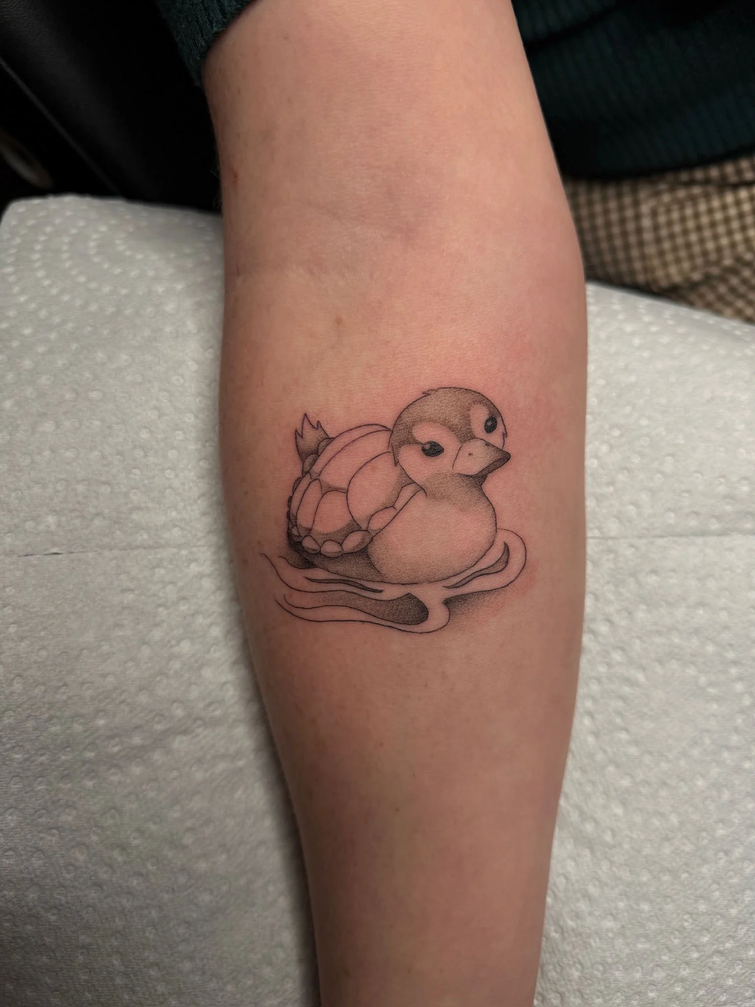 A tattoo of a cartoon-style duckling with a turtle shell on its back, floating on water.
