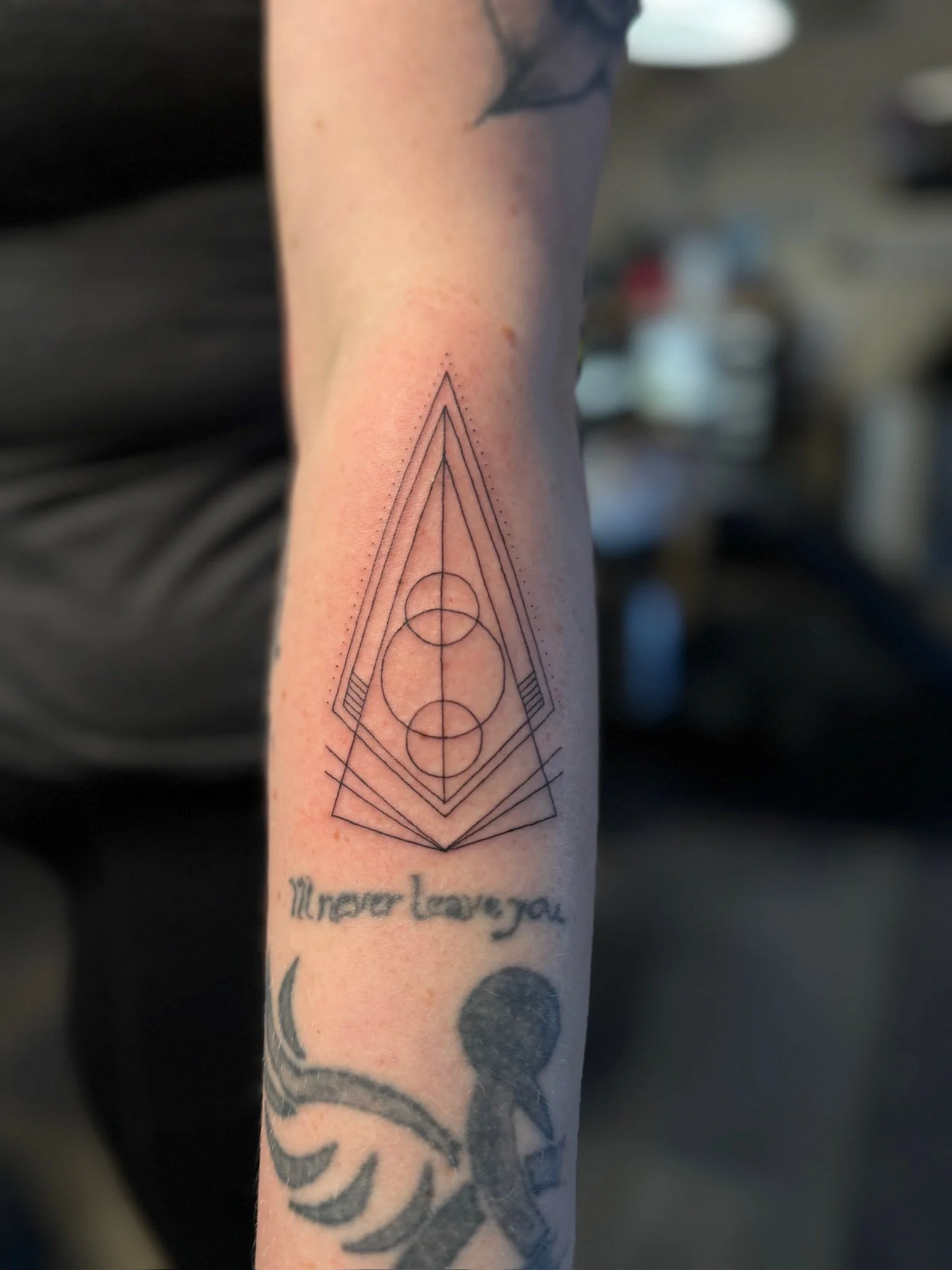 Tattoo of geometric shapes including circles and triangles on a person's forearm, with the words 'I never leave you' written below.