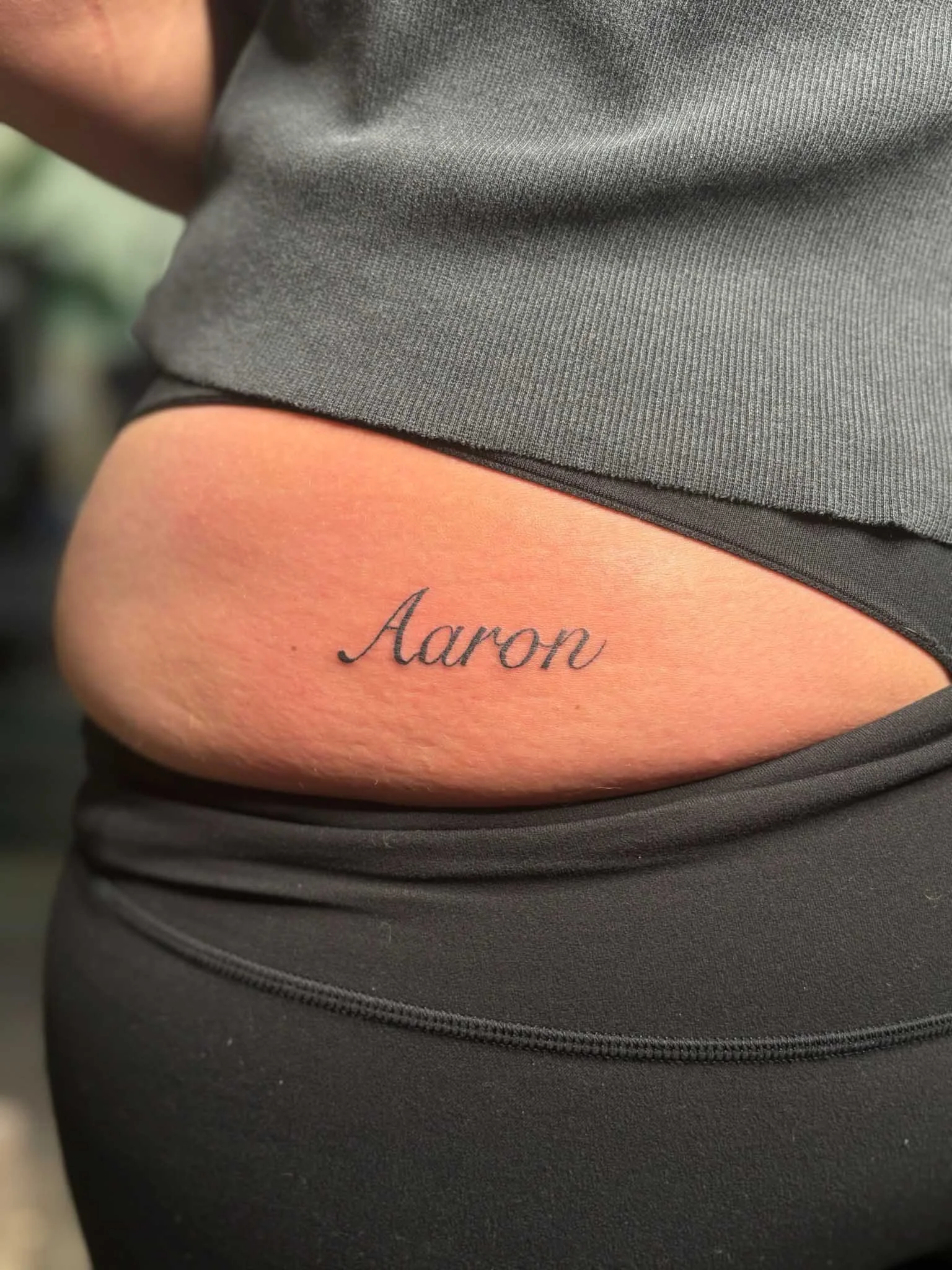 Tattoo of the name 'Aaron' on person's side near waist, with woman wearing gray athletic clothing.