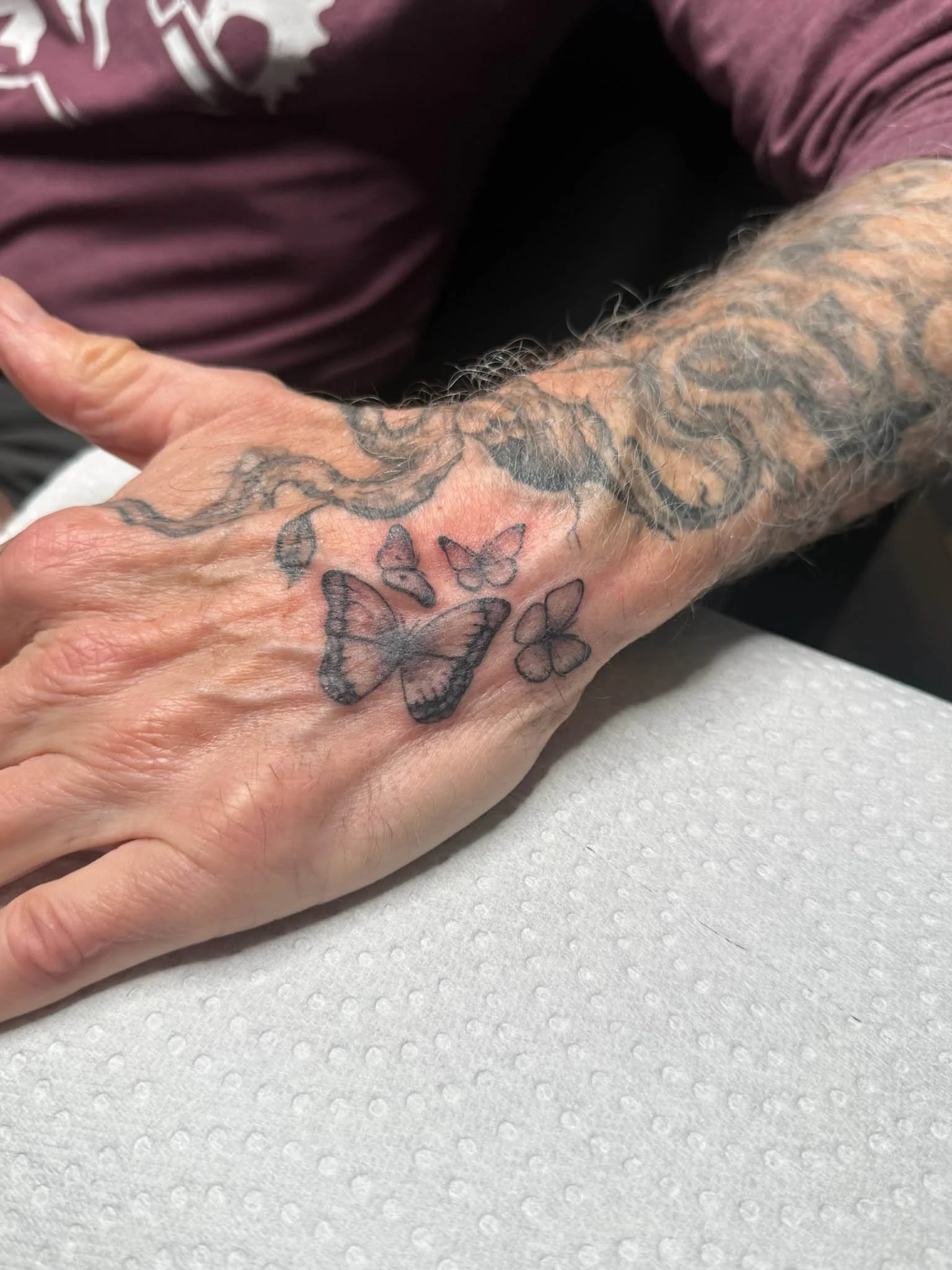 A person's hand with butterfly tattoos on the back, resting on a white textured surface, with arm tattoos visible.