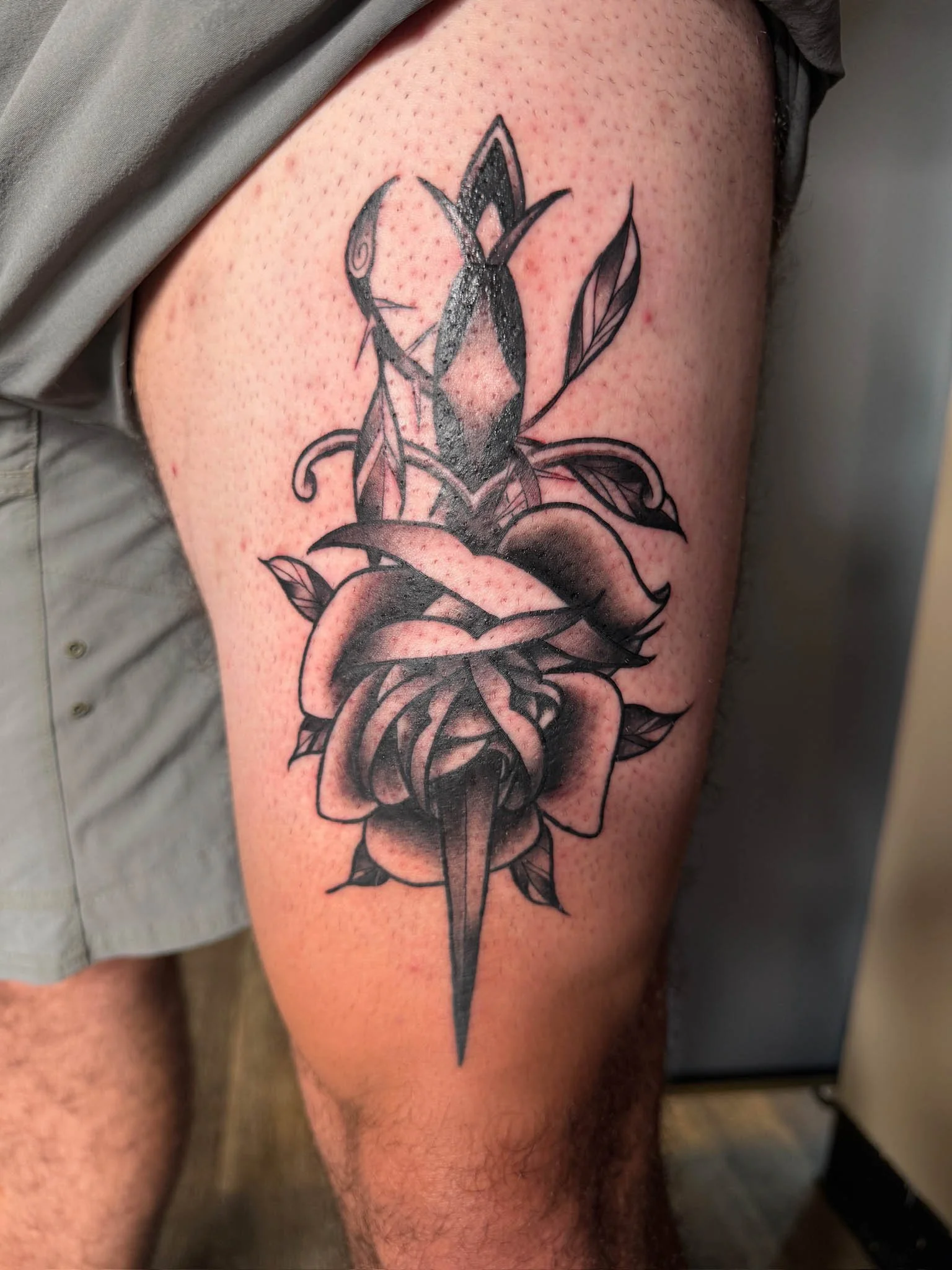 Black and gray tattoo of a sword piercing through a rose with leaves on a person's upper thigh.