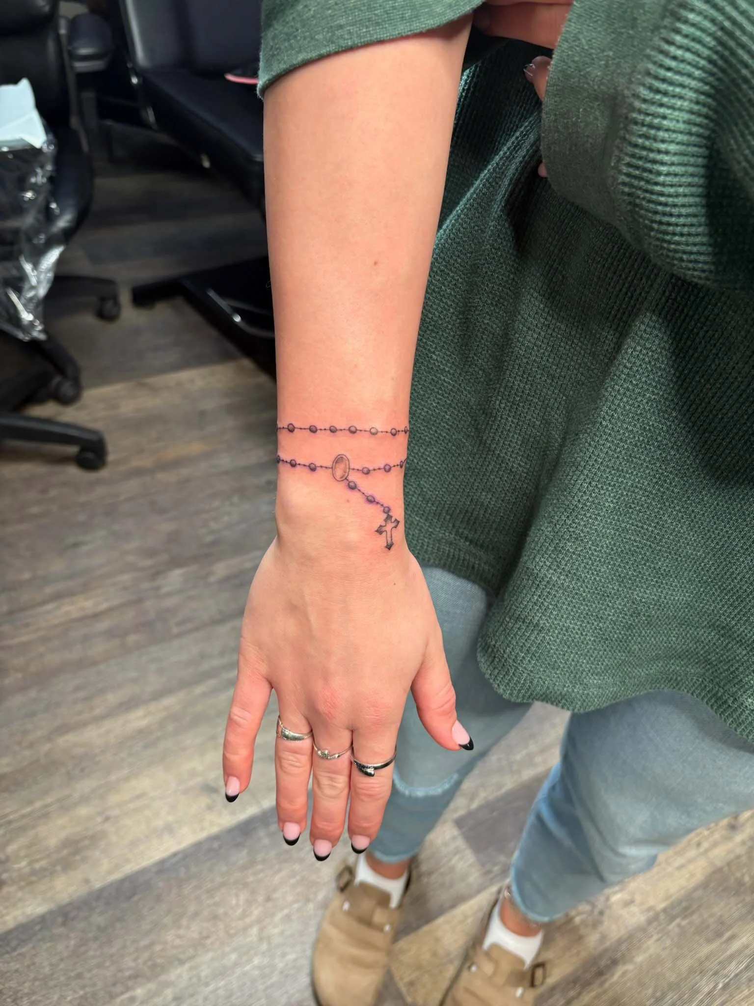 Close-up of a woman's left wrist with a delicate tattoo of a rosary, featuring beads, a cross, and a small heart-shaped medallion, in an office or indoor setting.