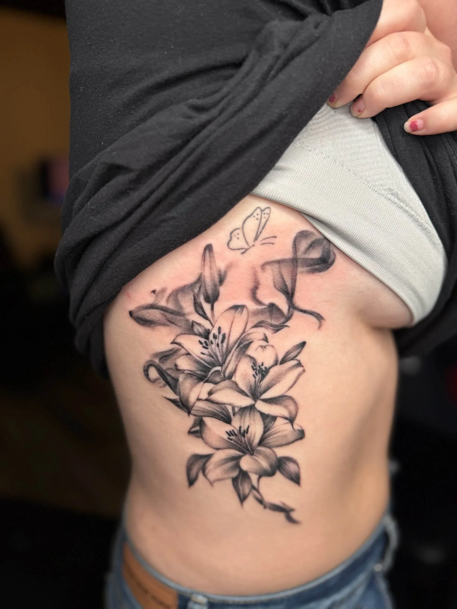 Black and gray floral tattoo of lilies on person's side, with butterflies above flowers.