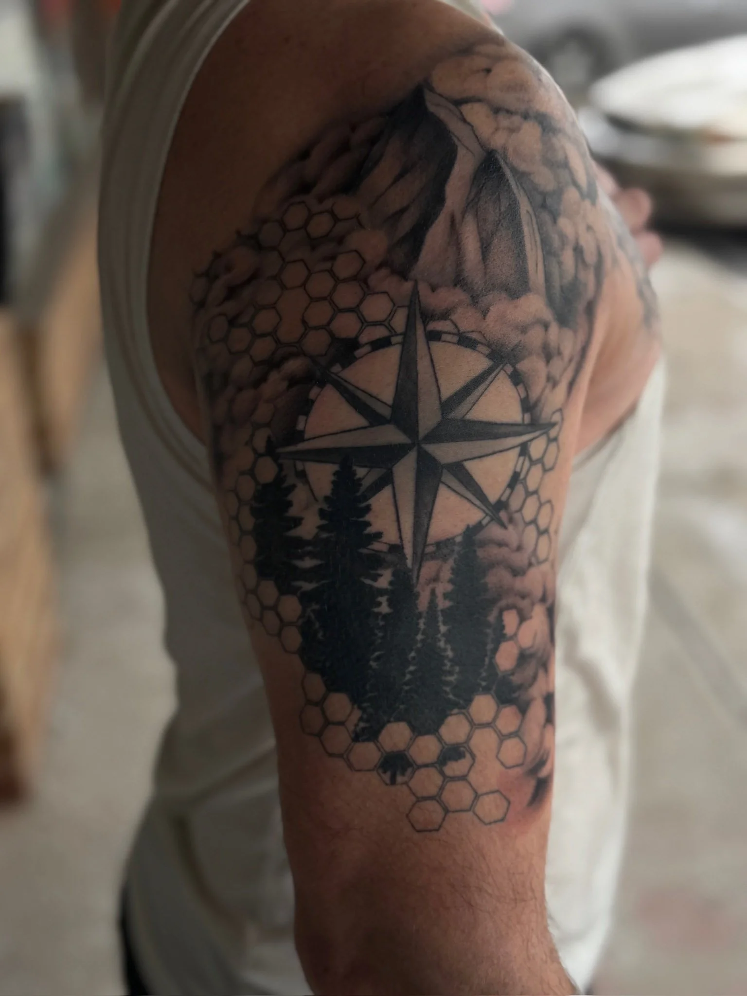 Tattoo of a compass rose, pine trees, honeycomb pattern, and clouds on person's upper arm.