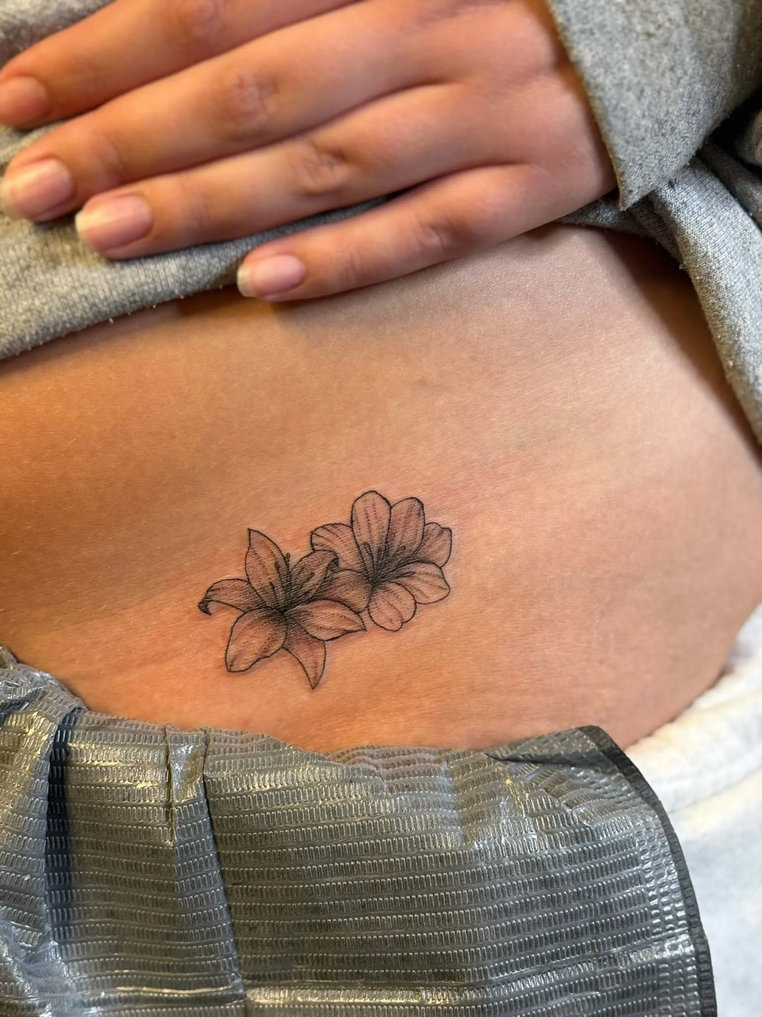 Close-up of a person's side with a tattoo of two lilies on their ribcage, partially covered by a gray sweatshirt.