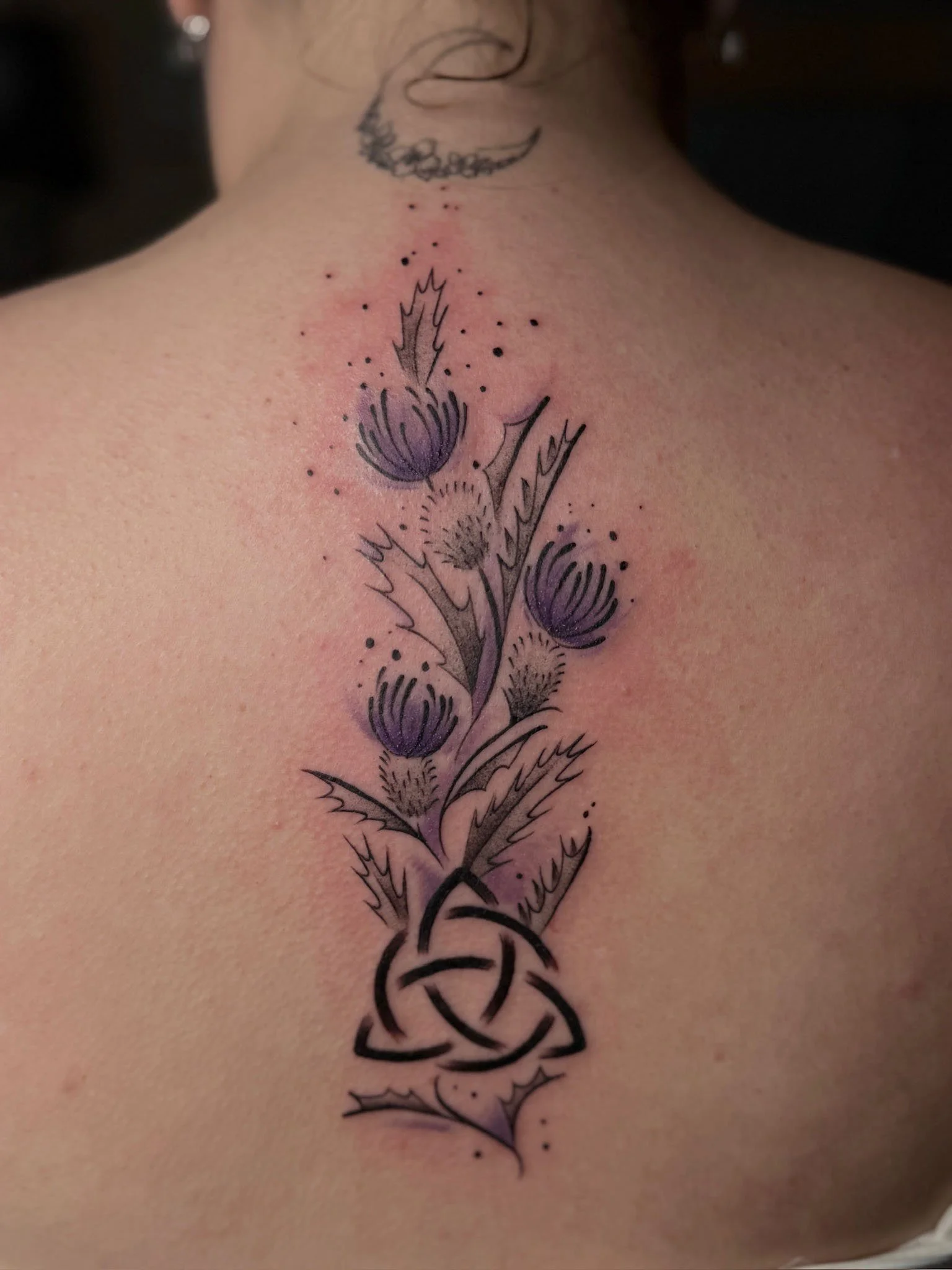 Tattoo on person's upper back featuring a floral design with three purple bud-like flowers, spiky leaves, and a black intricate knot at the bottom.