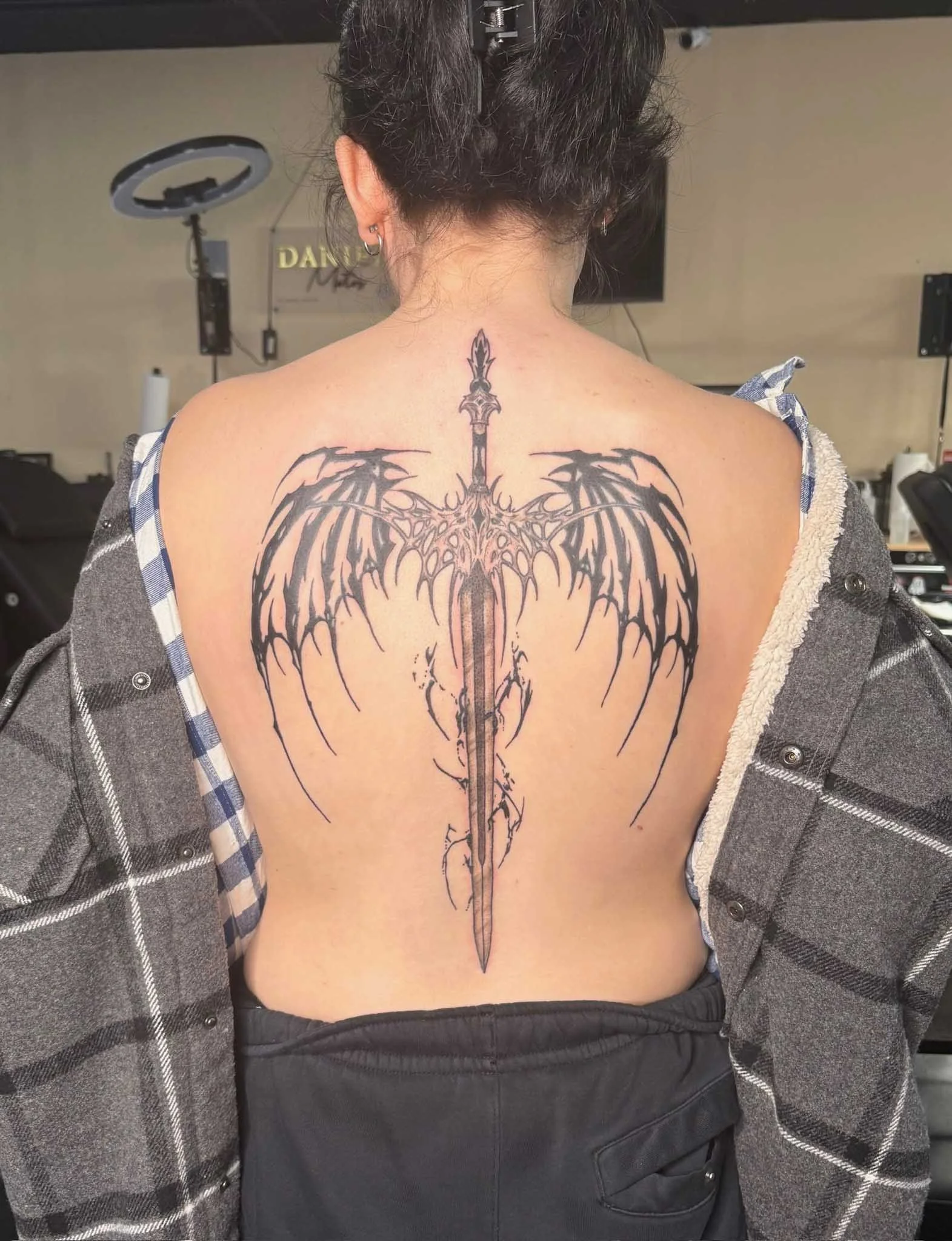 Tattoo of a dark, ornate sword with outspread wings on a person's back.