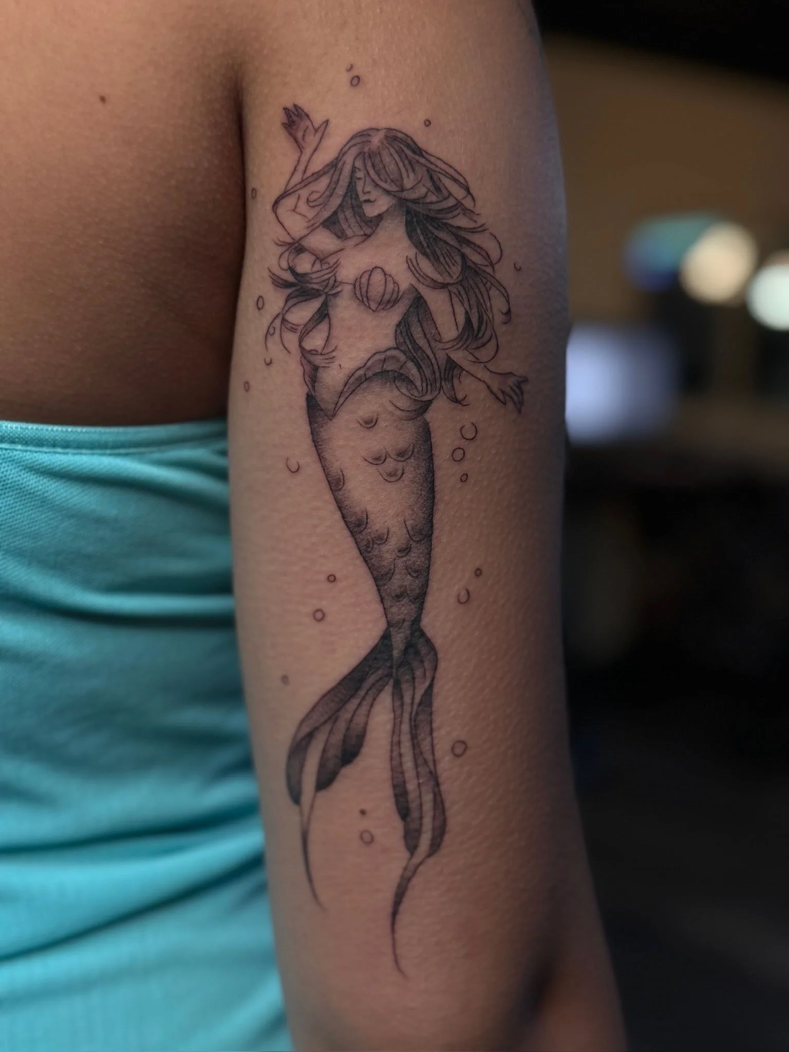Tattoo of a mermaid with flowing hair, holding a shell, and surrounded by bubbles on person's upper arm.
