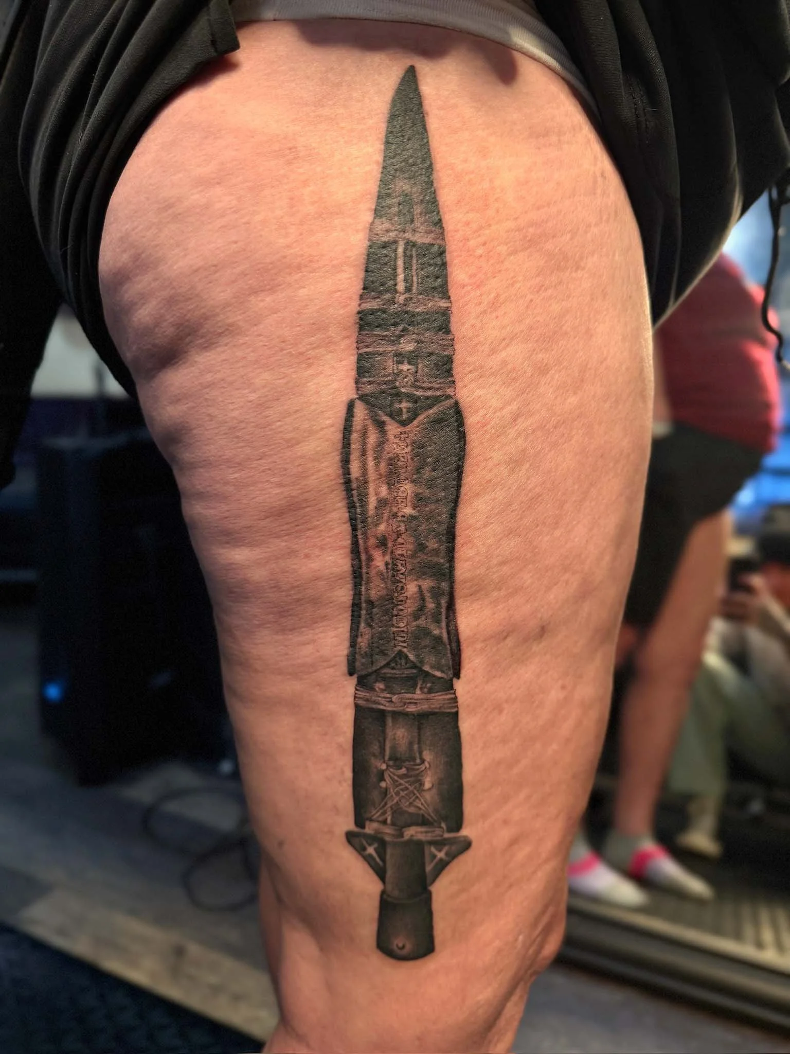 A tattoo of a large combat knife on a person's thigh, featuring a detailed blade with inscriptions and a handle with decorative elements.