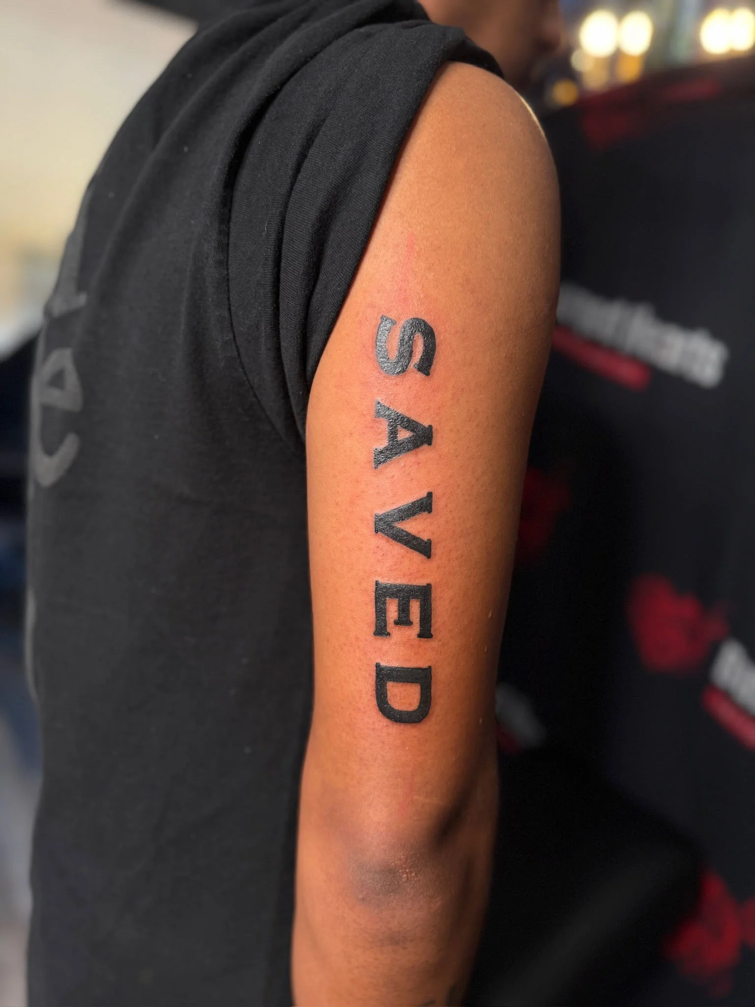 Tattoo on person's upper arm reads 'SAVED' in black ink.