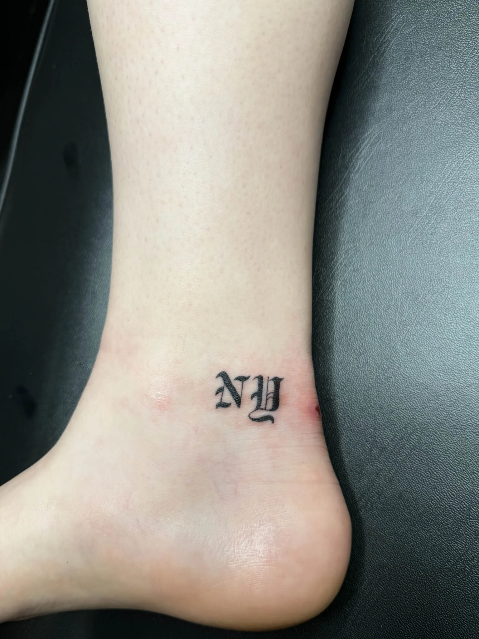 Tattoo of the letters 'NY' in black ink on the ankle.