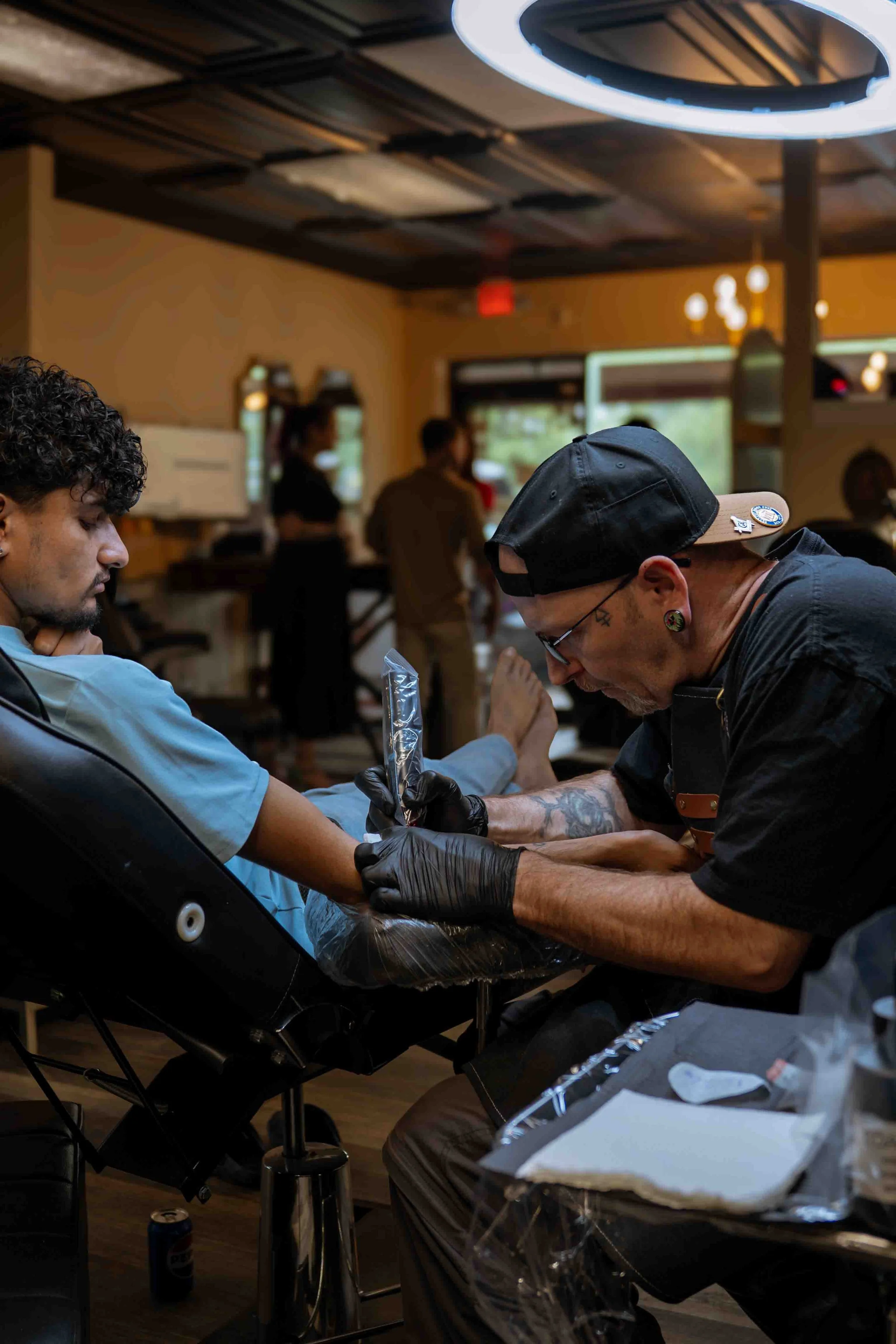 A man receiving a tattoo on his arm from a tattoo artist in a tattoo parlor. The artist is wearing gloves and a hat, focused on his work. The background shows other people in the parlor and tattoo equipment.