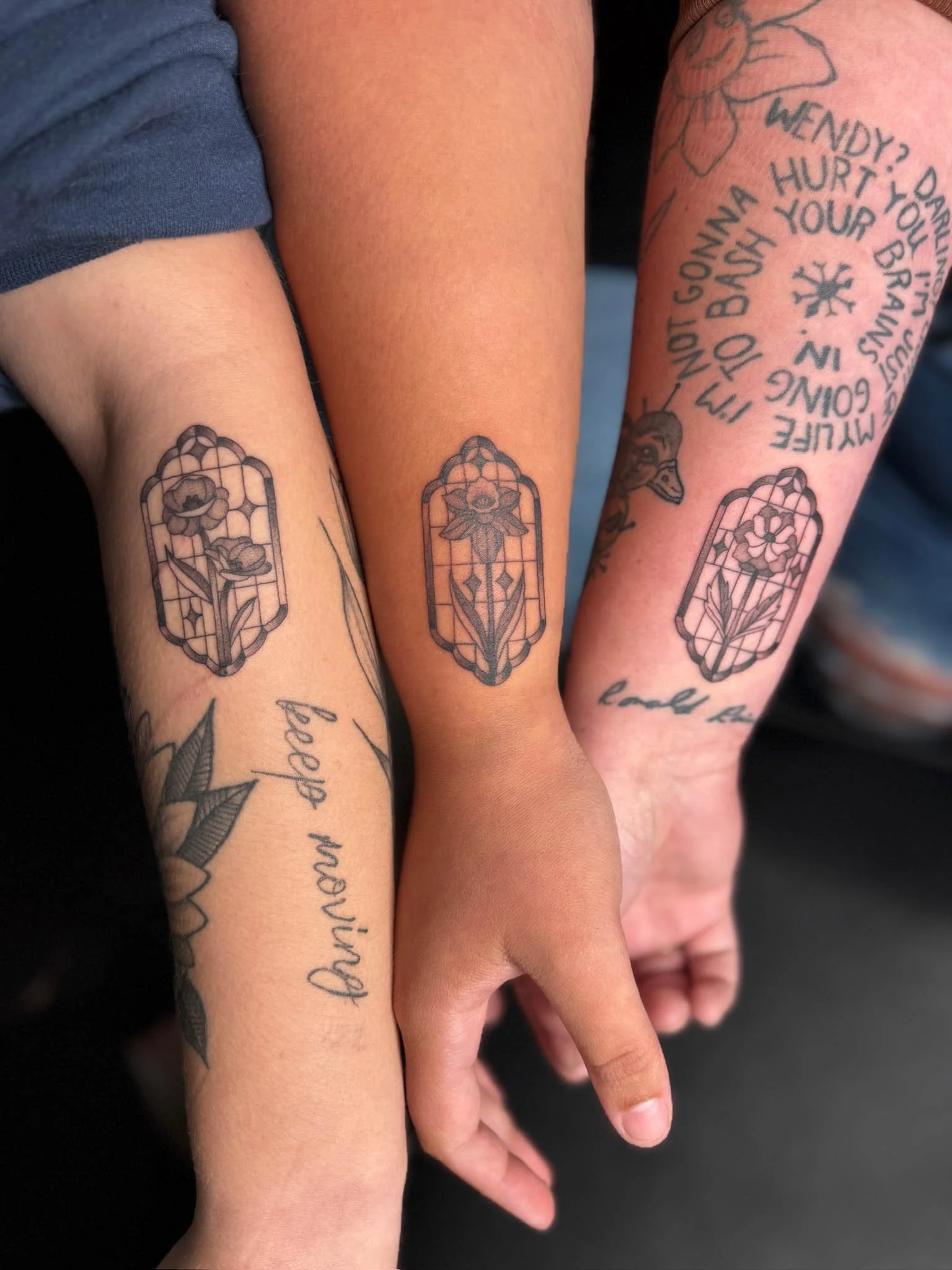 Three arms with tattoos, each featuring a stained glass window design with a flower. One arm has a tattoo of a quote in cursive reading 'Dreams Doork'.
