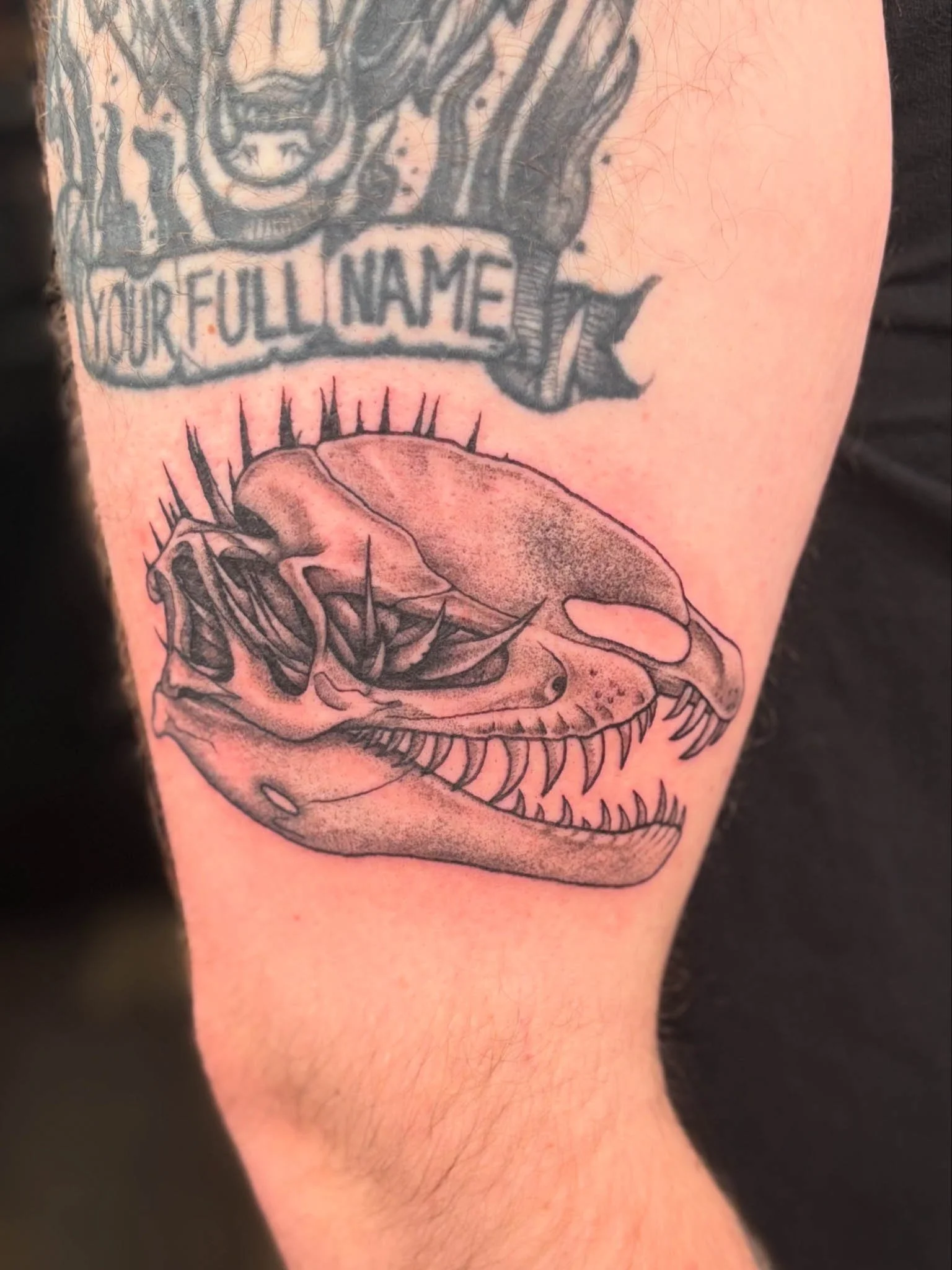 Tattoo of a dinosaur skull with sharp teeth, detailed shading, and a banner above it with the words "Your Full Name".