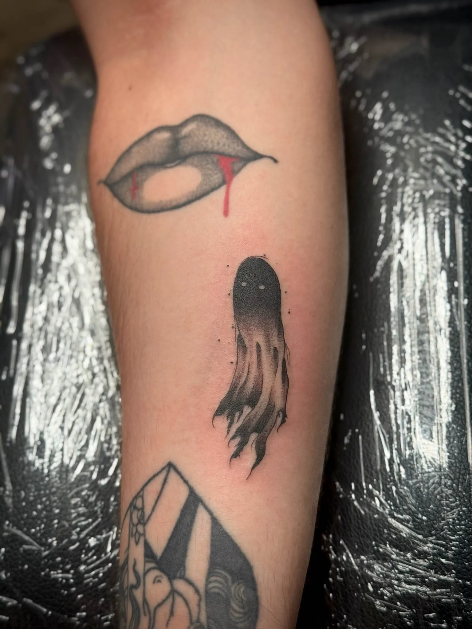 Tattoo of lips with a droplet of red hanging from the lower lip and a small ghost-like figure beneath, with part of a geometric design visible below.
