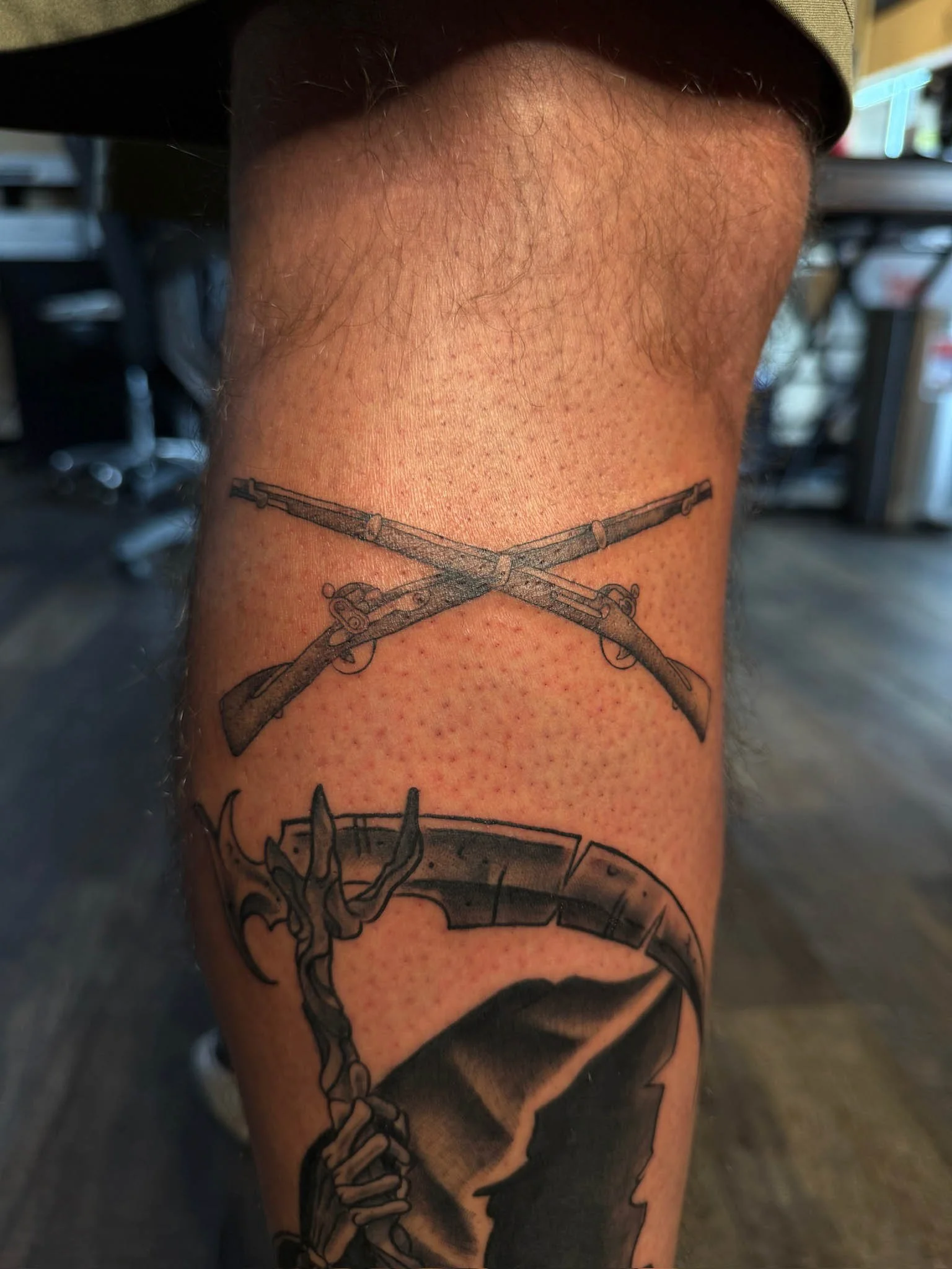 Tattoo of crossed guns above a scythe with a skeletal hand gripping the handle on a person's leg.