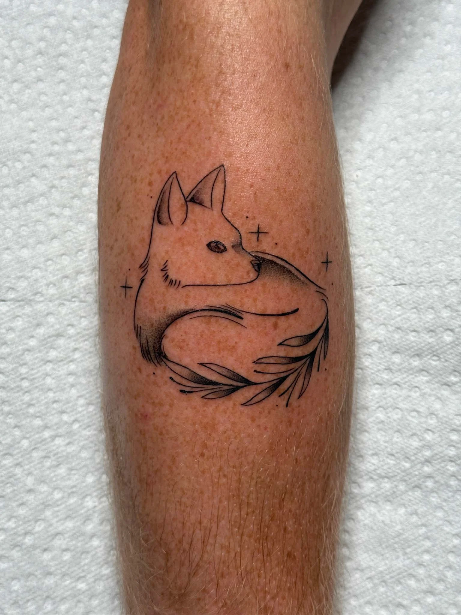 Minimalist tattoo of a fox curled up with eyes closed, surrounded by small stars and leaves.