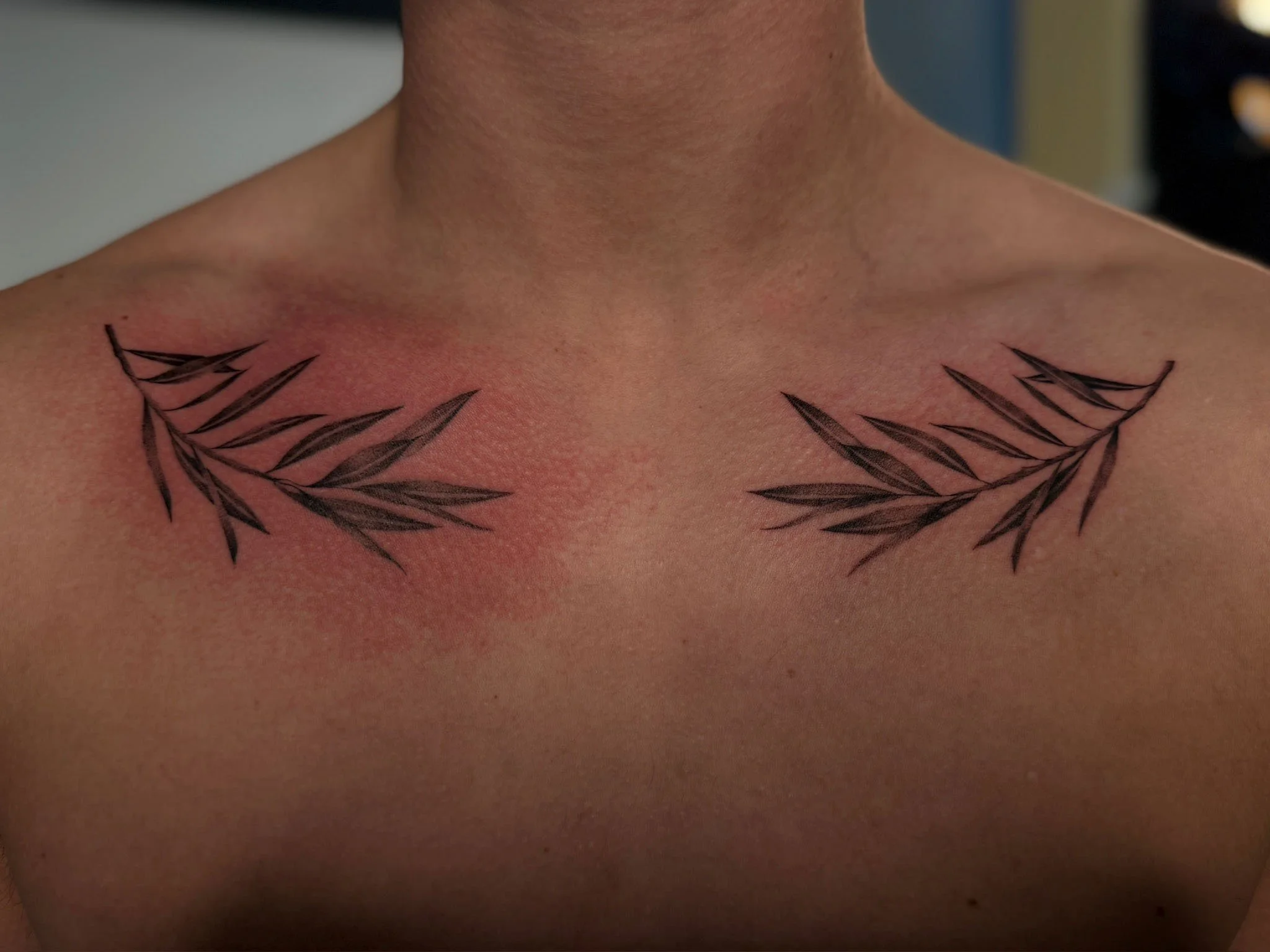 Tattoo of two black leaf branches on the upper chest, near the collarbones.