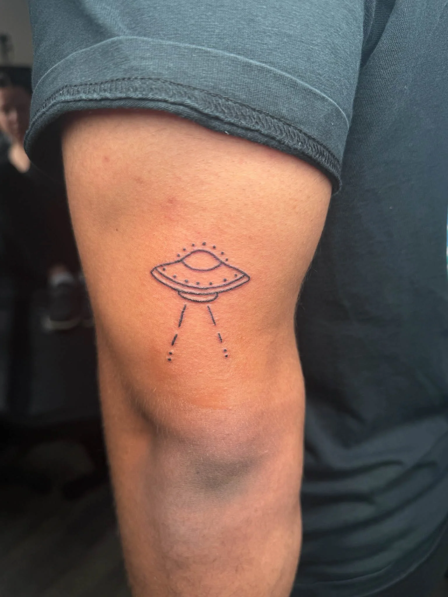Tattoo of a flying saucer on a person's upper arm, depicted with dotted lines and rays emanating from it.