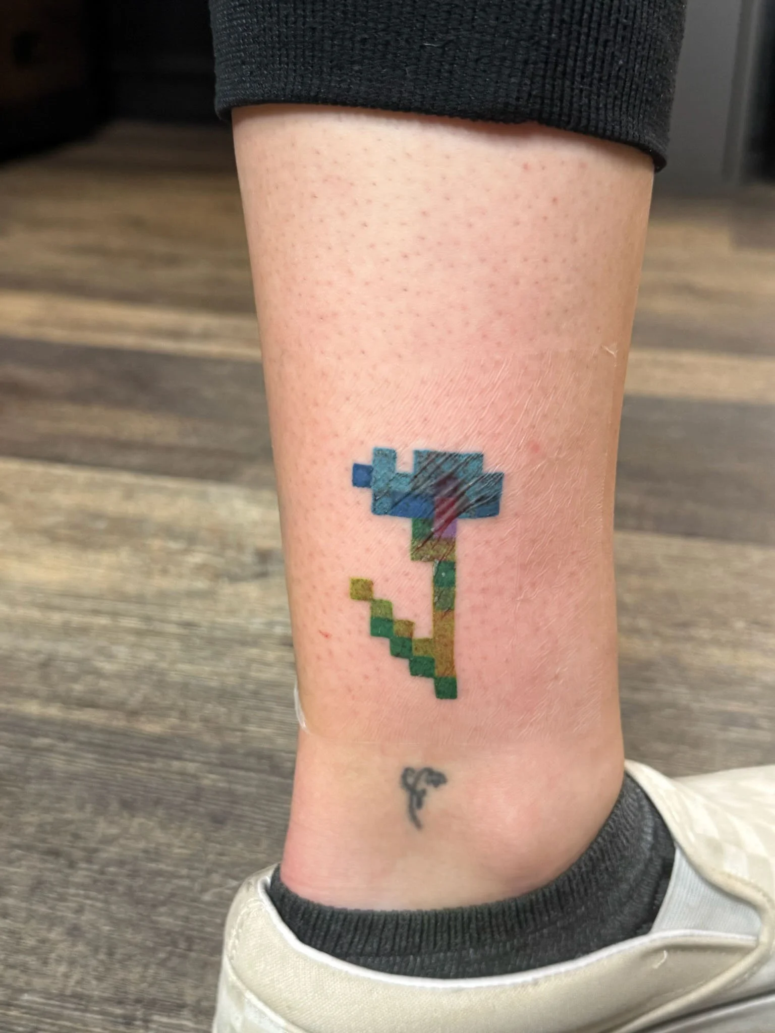 A tattoo of a pixel art flower on a person's ankle, with a small black symbol below it.