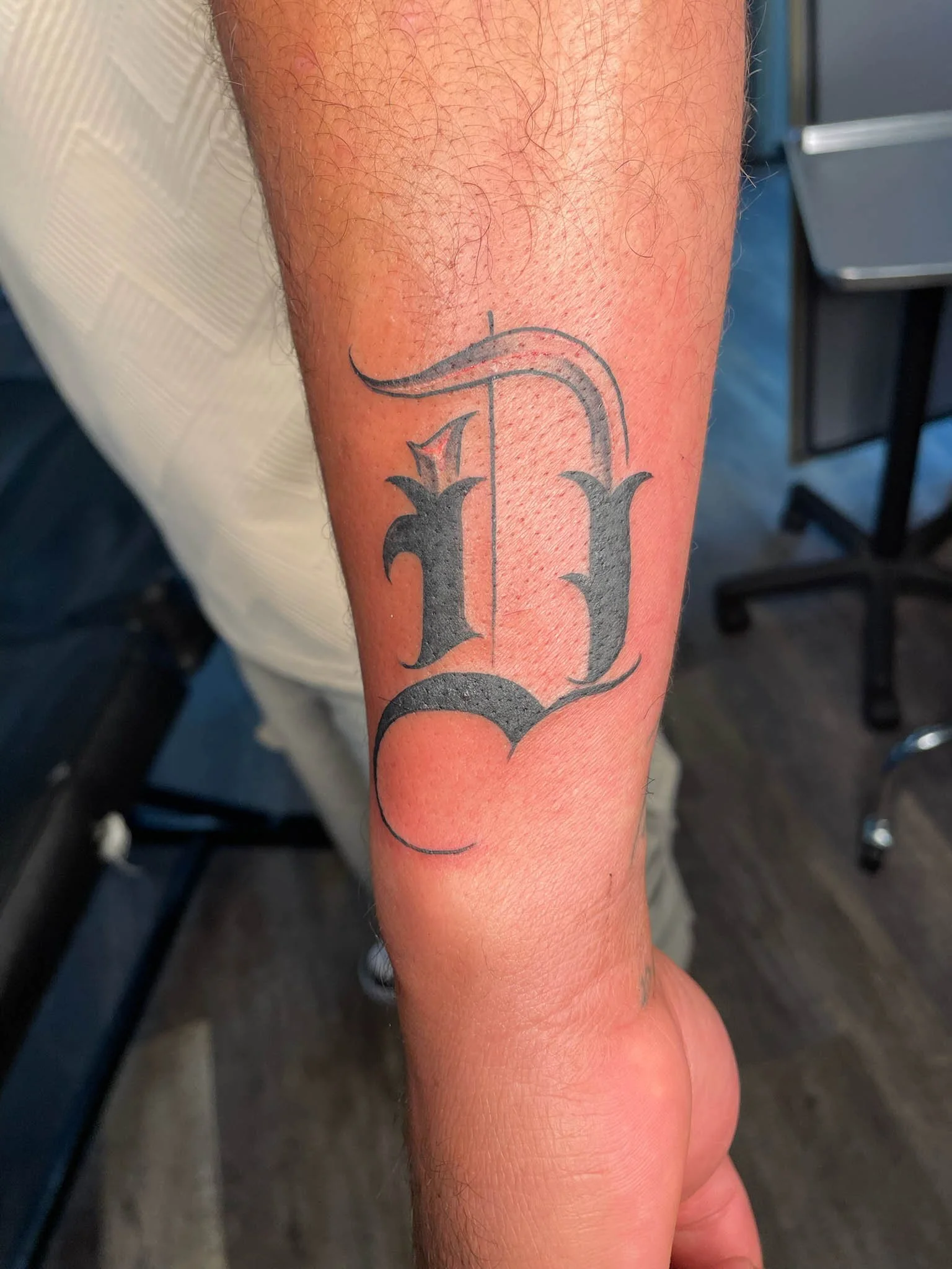 A person's forearm with a fresh tattoo of the large, black letter 'L' in a Gothic style font.
