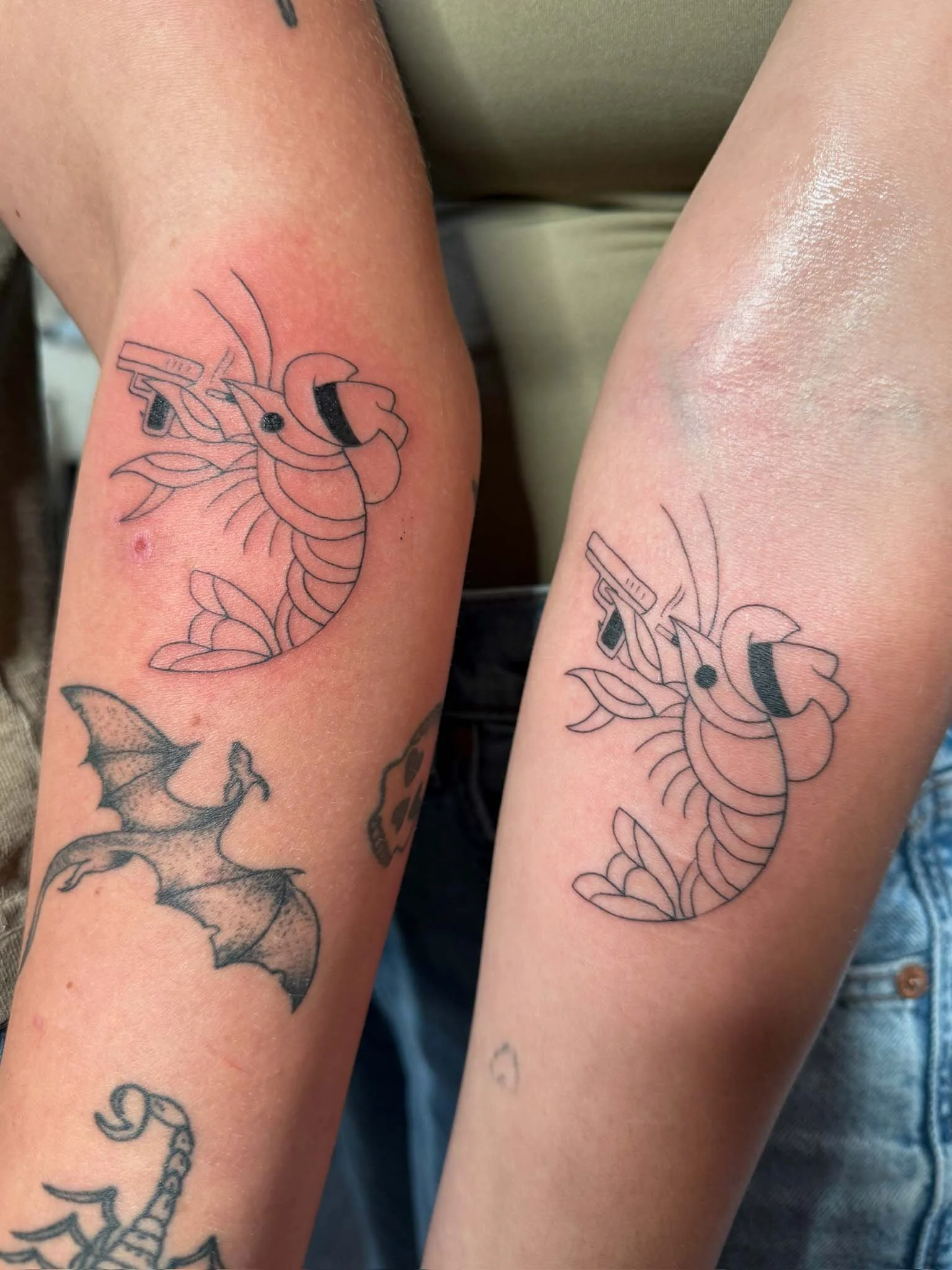 Two arms with matching black line tattoos of a shrimp holding a gun and a bat with wings, and other tattoos of a bat and a cartoonish skull.