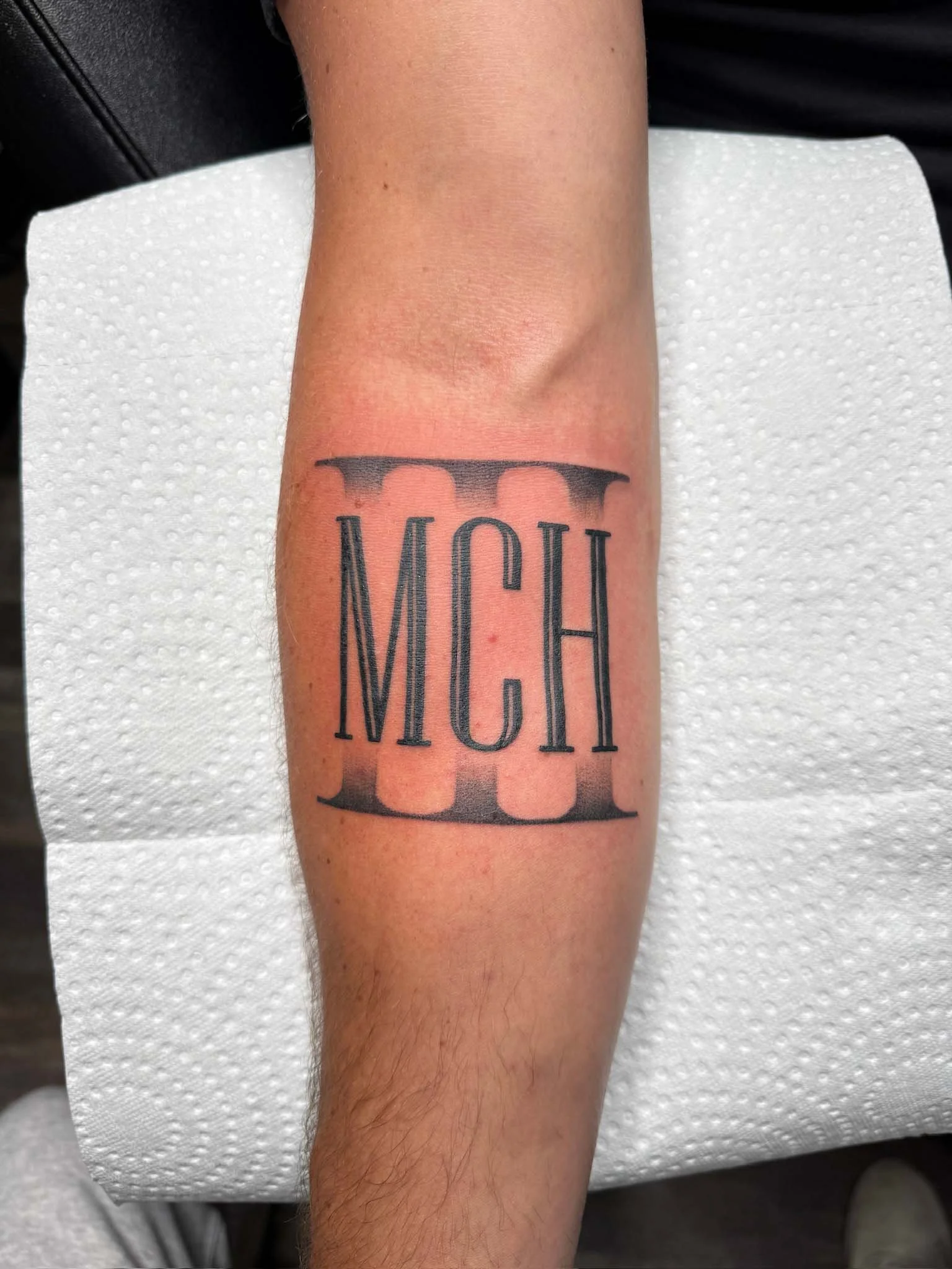 Tattoo of the letters 'MCH' with a black tribal-style band above and below, on a person's forearm.