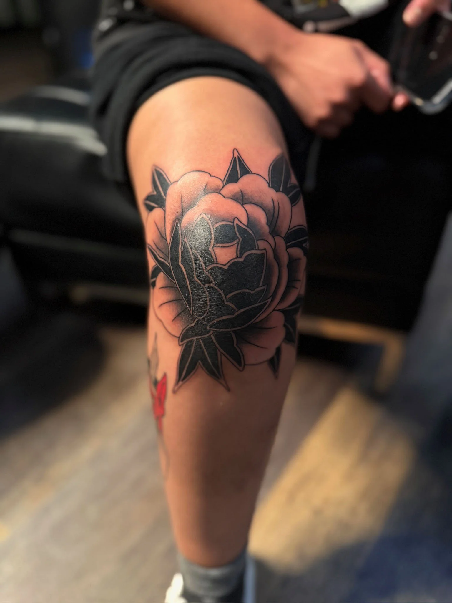 A person with a large black and gray floral tattoo on their thigh, featuring a blooming flower with detailed petals and leaves, sitting on a black chair.