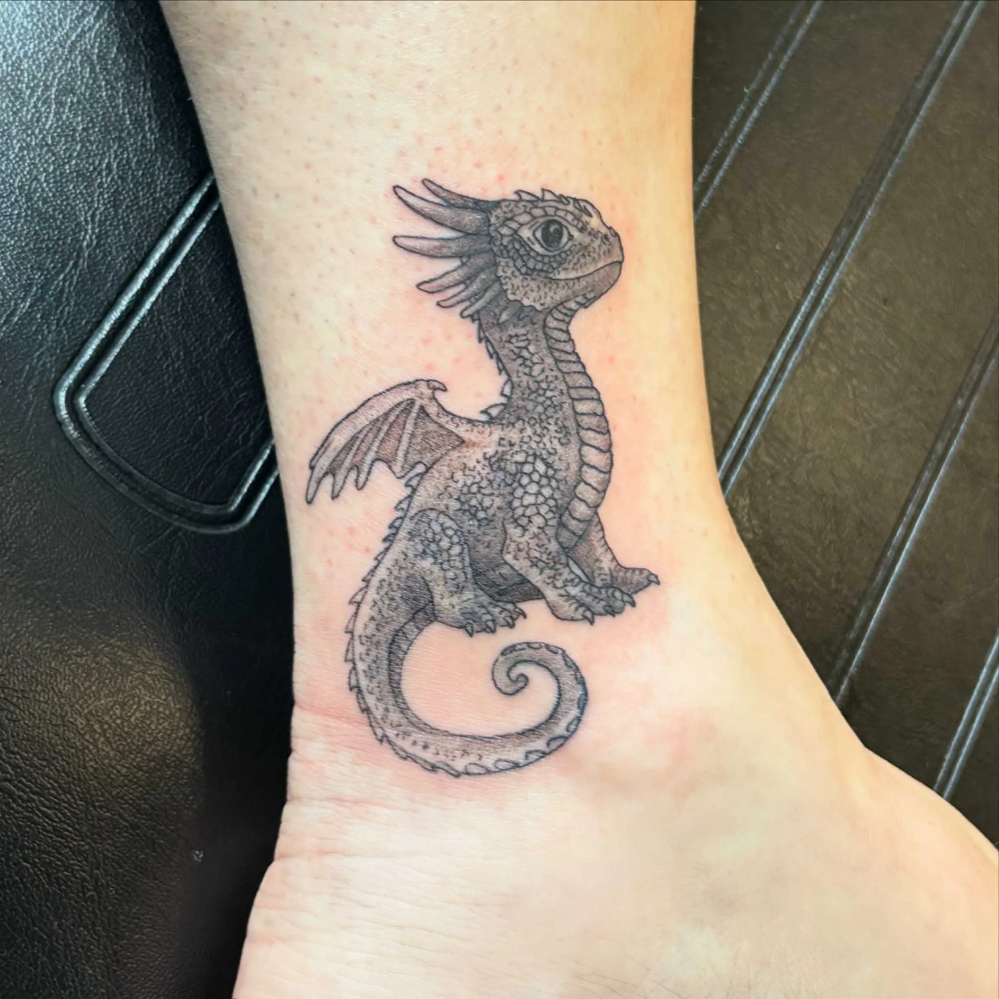 Black and gray tattoo of a baby dragon with detailed scales, wings, and a curled tail, on a person's ankle.