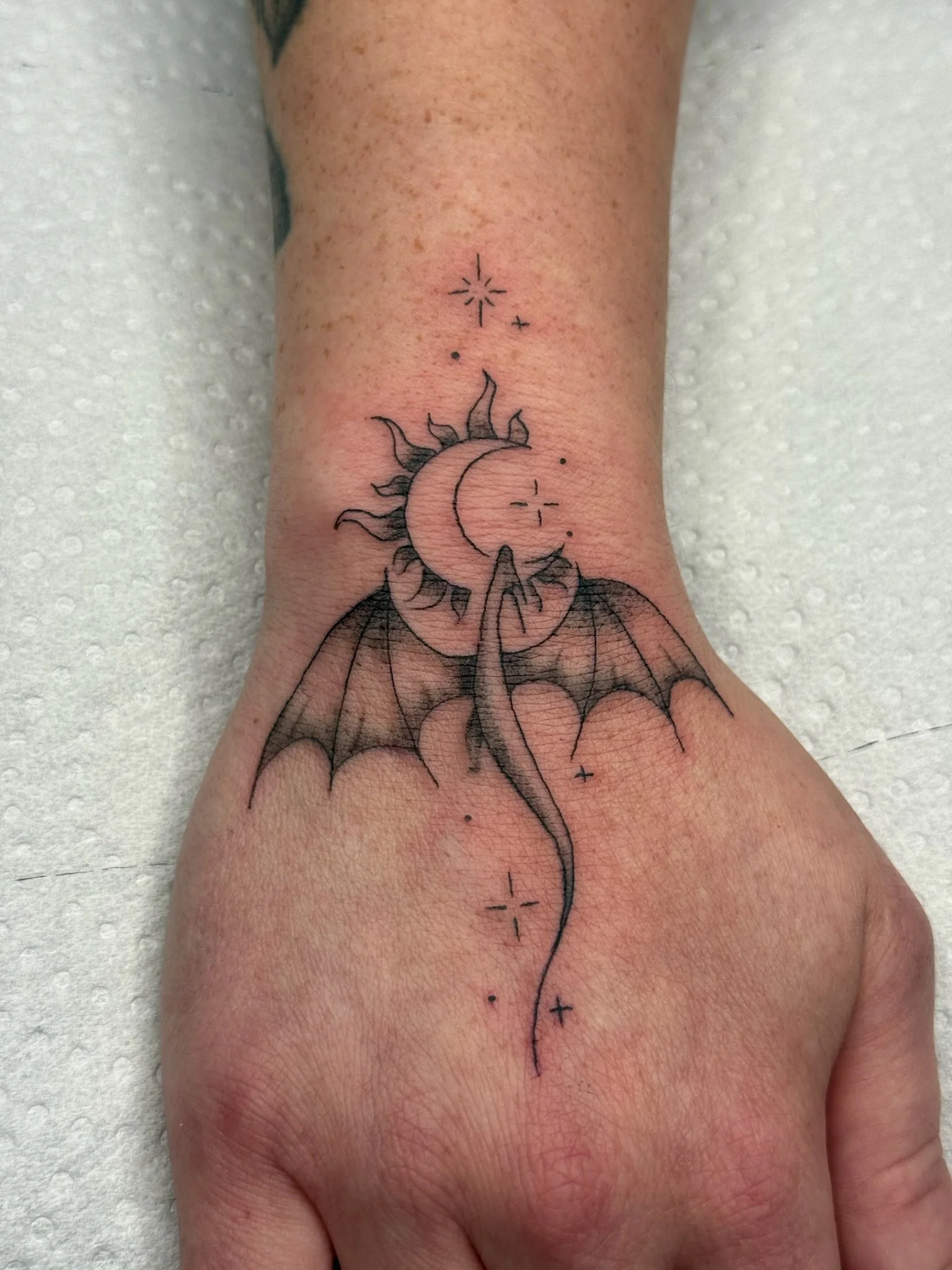 Tattoo of a crescent moon with bat wings, surrounded by stars and dots, on the person's forearm.