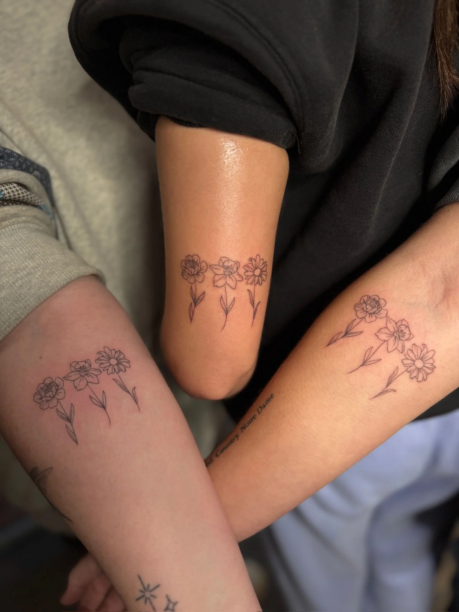 Three arms with matching floral tattoos featuring daisy-like flowers and leaves.