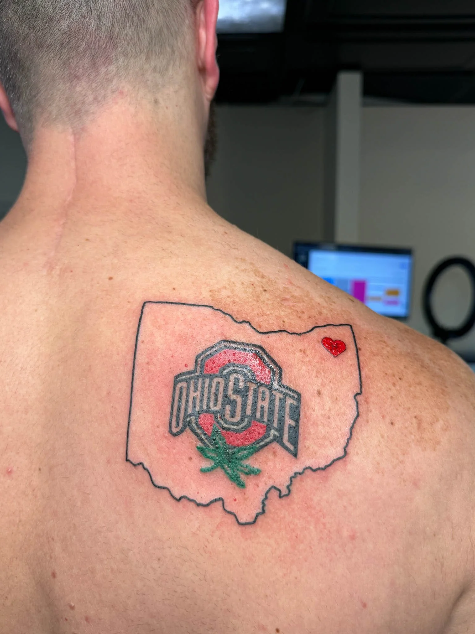 Tattoo on person's upper back featuring the Ohio State logo within an outline of the state of Ohio, with a small red heart above it.
