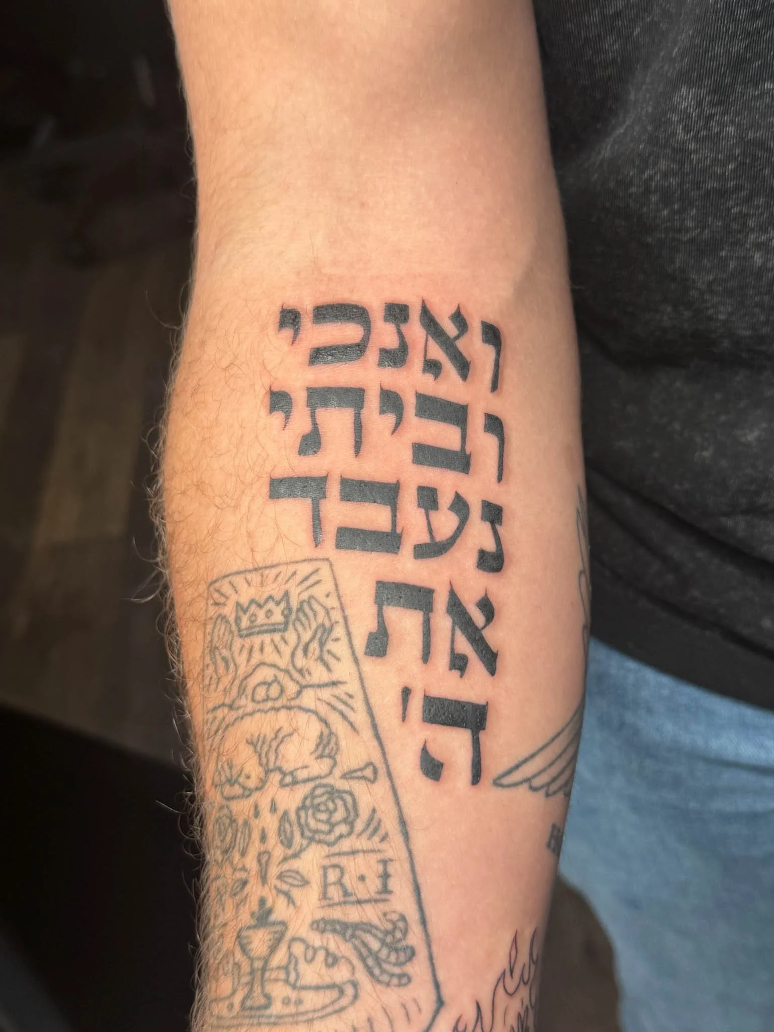 Tattoo of a Hebrew phrase and a partially visible design with a crown, a lion, a rose, and other symbols on a person's forearm.