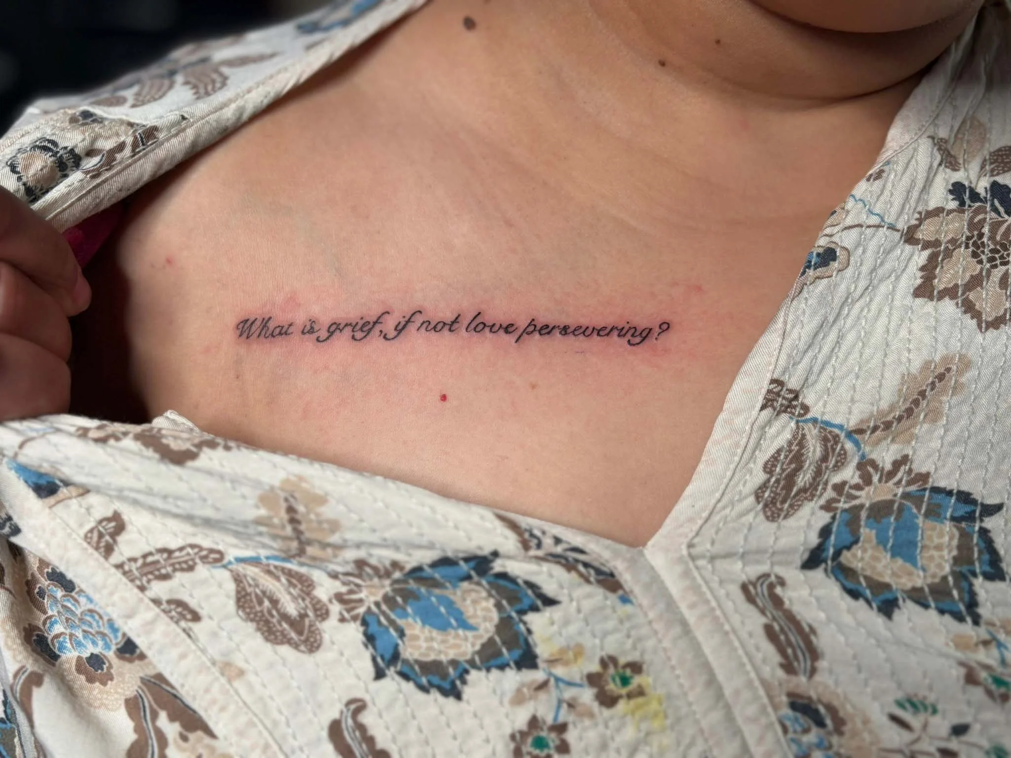 A tattoo on a person's chest reads, 'What is grief, if not love persevering?' The person is wearing a floral-patterned shirt partially unbuttoned.