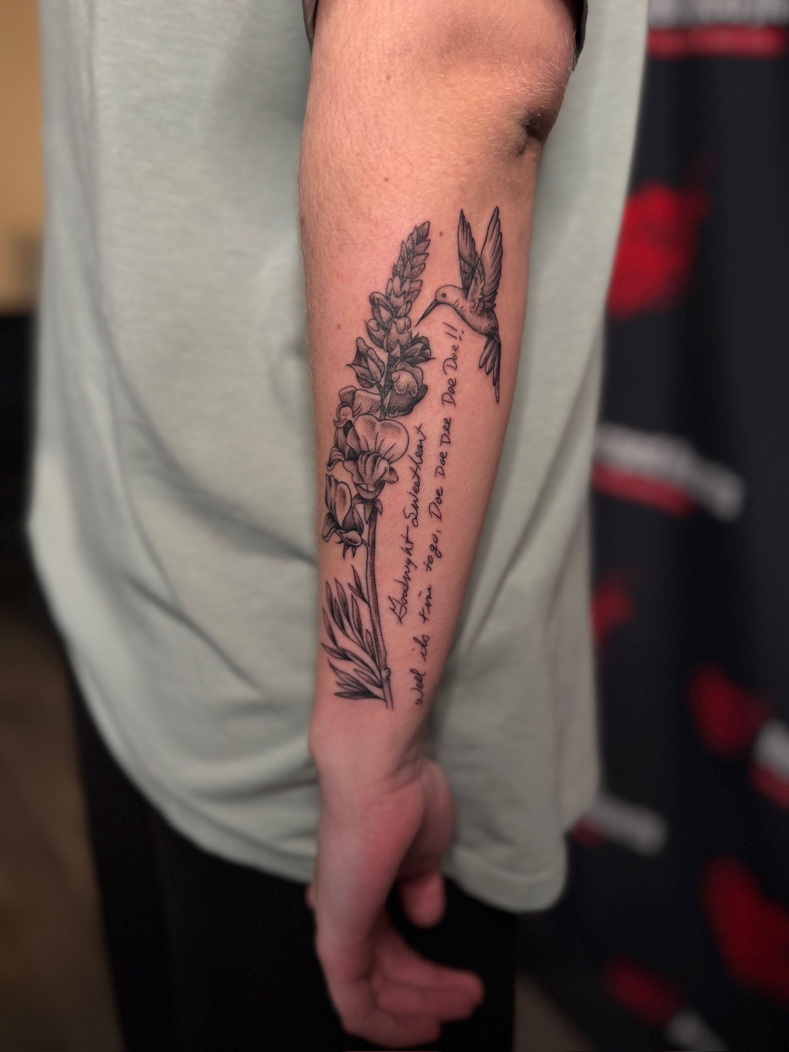 Black and gray tattoo of a hummingbird and a tall flower with handwritten-style script, on the inner forearm of a person wearing a gray shirt.