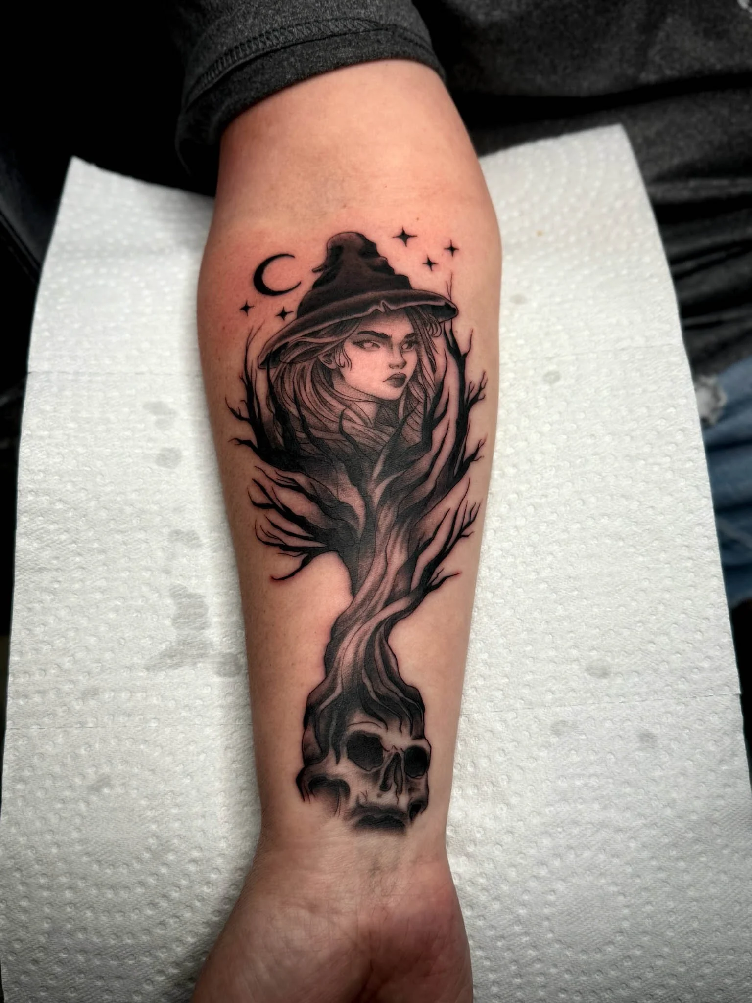 Tattoo of a witch wearing a hat with a moon and stars above, a woman's face in the hair, tree branches, and a skull at the bottom.