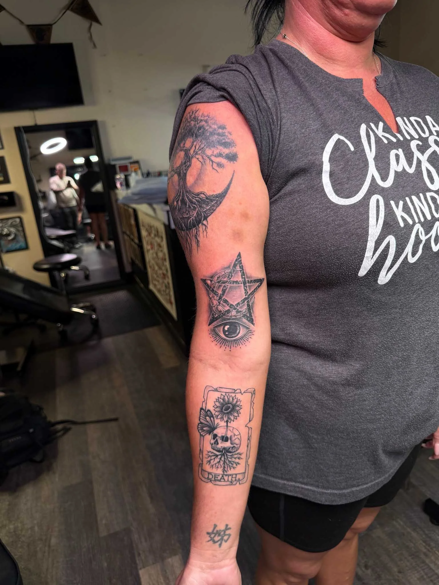Black and gray arm sleeve tattoos including a tree of life inside a crescent moon on the shoulder, a pentagram over an eye above the elbow, a "DEATH" tarot card with a skull and sunflower on the forearm, and a small kanji character on the wrist