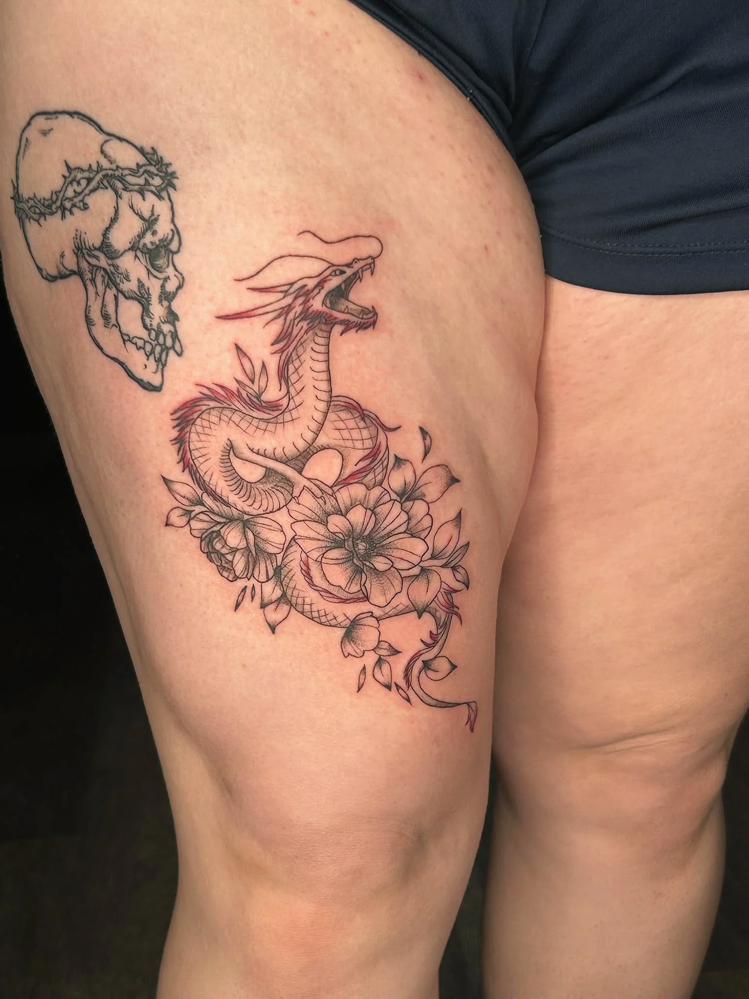 Tattoo of a dragon with flowers on a person's upper thigh.