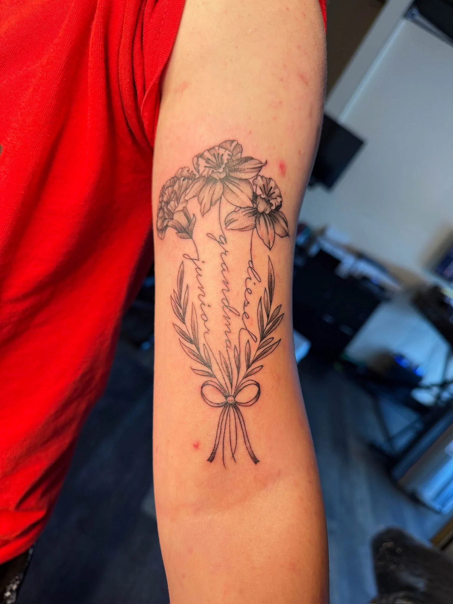 A black line tattoo of a bouquet of flowers with a bow at the base, with handwritten names 'Victoria,' 'Hamdab,' and 'Huni' in cursive script.