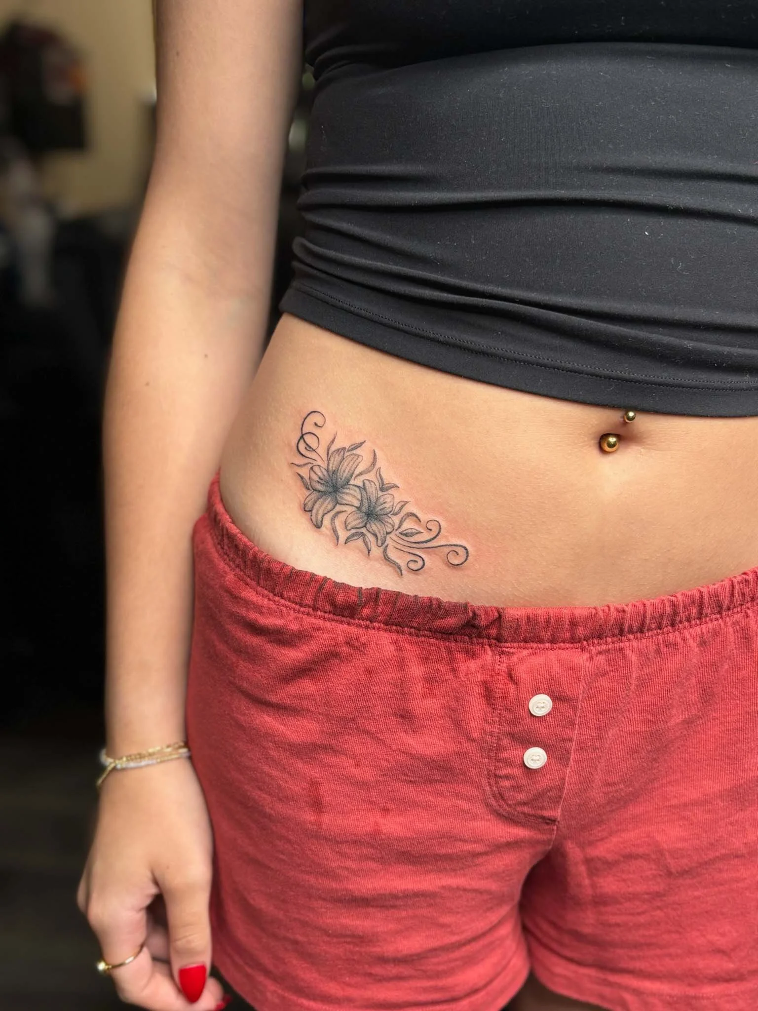 Close-up of a woman's midsection showing a floral tattoo, and a navel piercing, wearing a black crop top and red shorts.