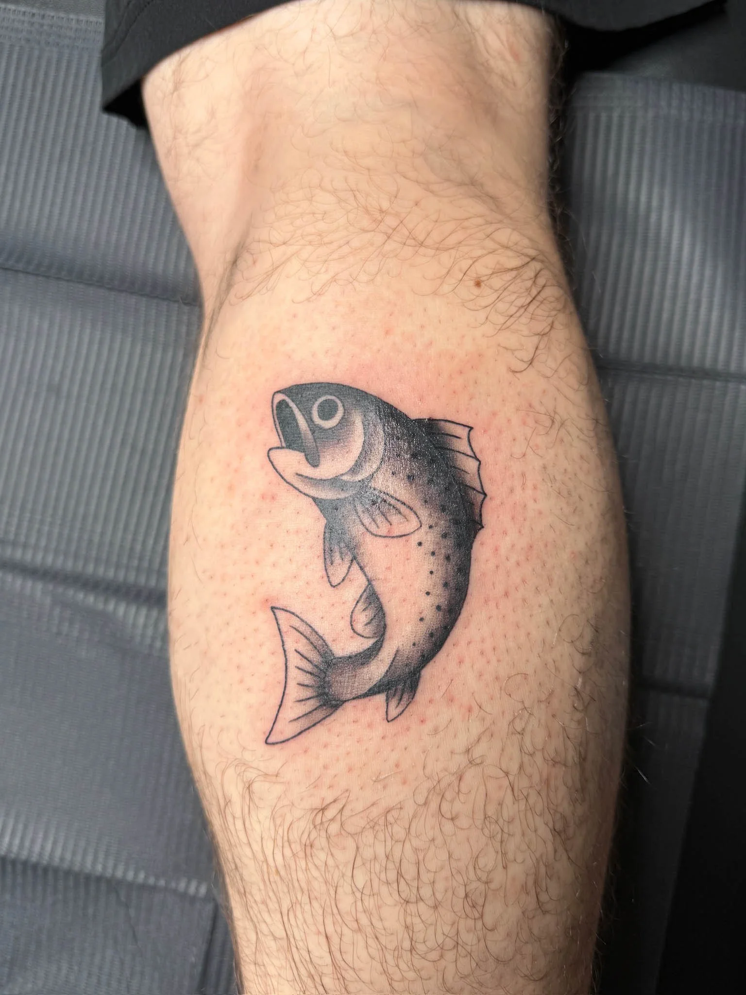 Black and gray tattoo of a fish on a person's leg, with the fish depicted in a realistic style with detailed scales and fins.