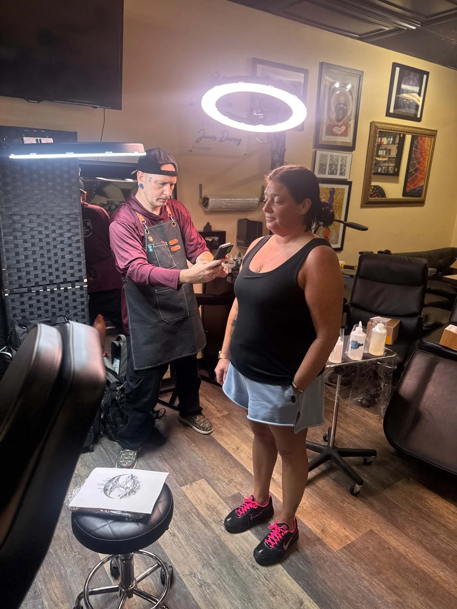 Tattoo artist wearing an apron taking a photo with his smartphone of a client standing inside a studio