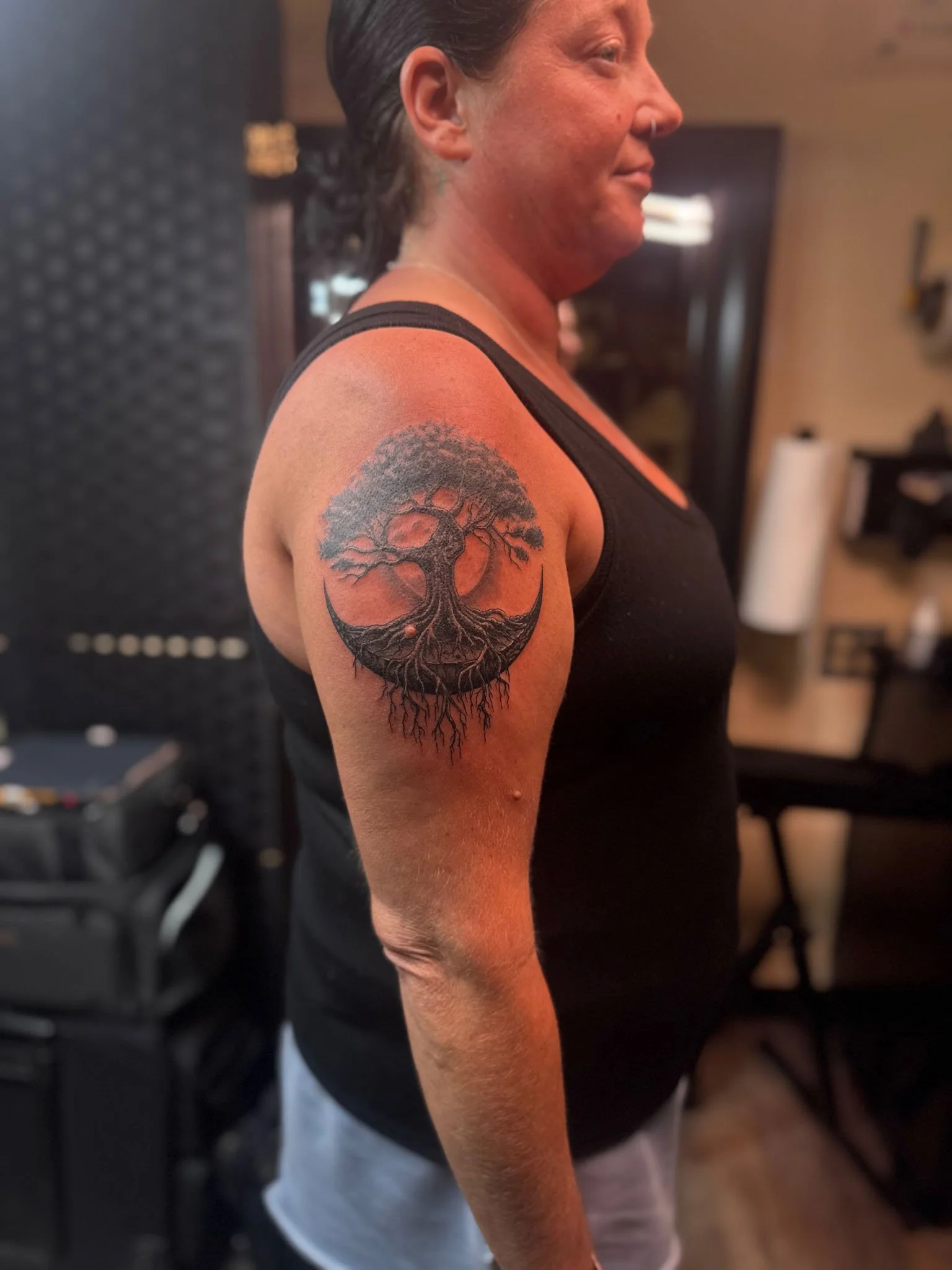 Black and gray tattoo of a gnarled tree of life with its roots interlaced inside a crescent moon shape, placed on a woman's upper arm.