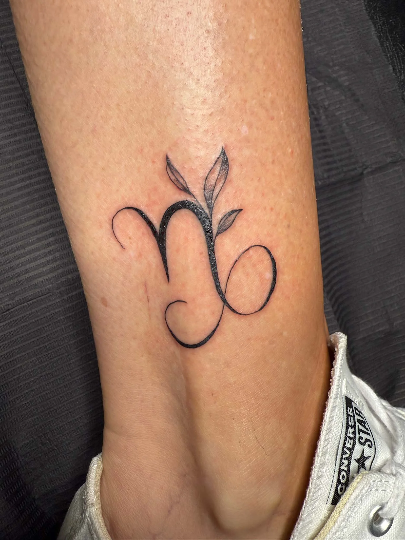 A tattoo on a person's forearm featuring a black cursive letter 'N' that is a Zodiac symbol with three leaves above it.