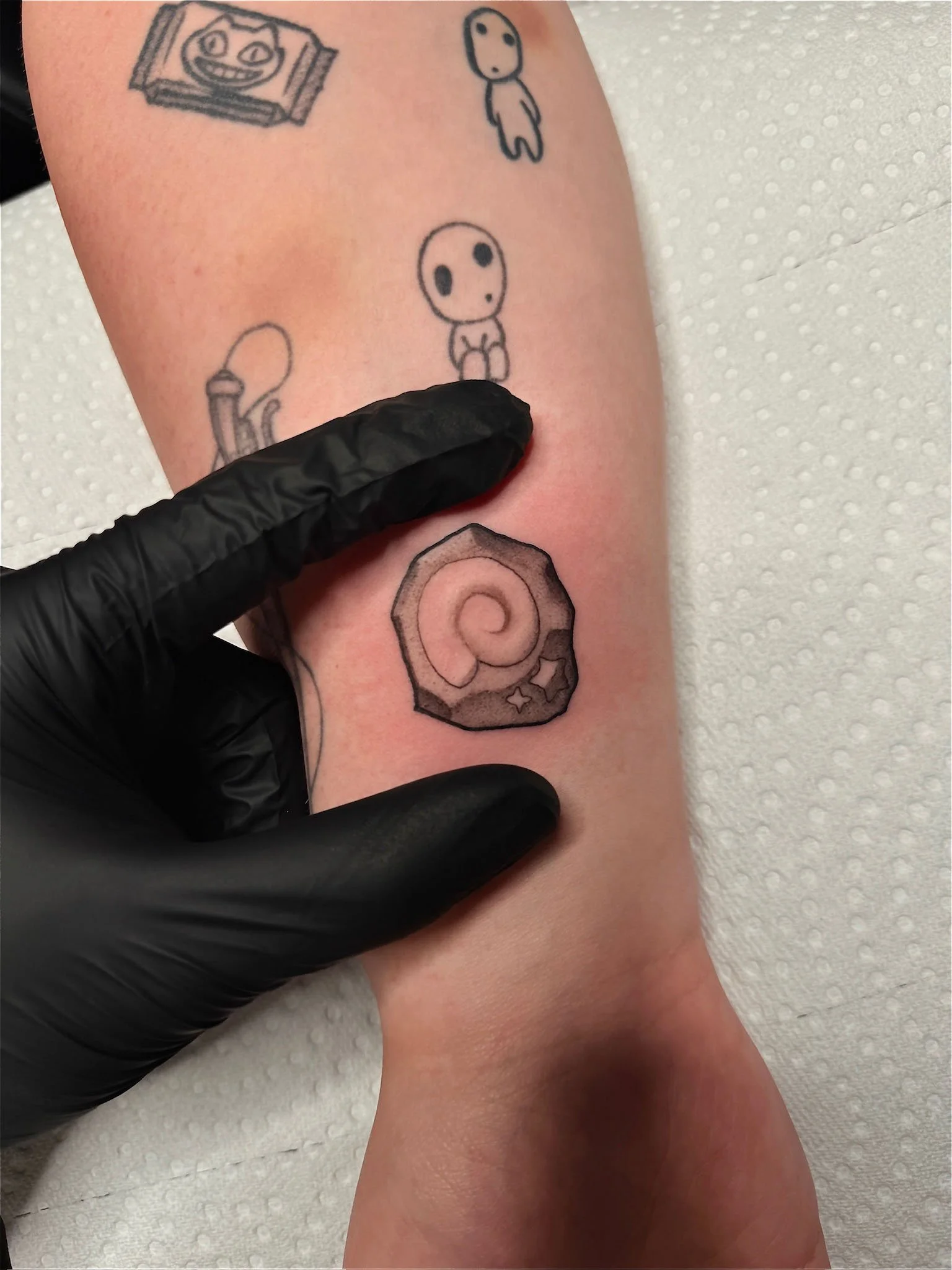 Tattoo artist applying a tattoo of a spiral shell on a person's arm. The tattoo is done in black and grey with some shading, and a small star and cross are visible near the shell.